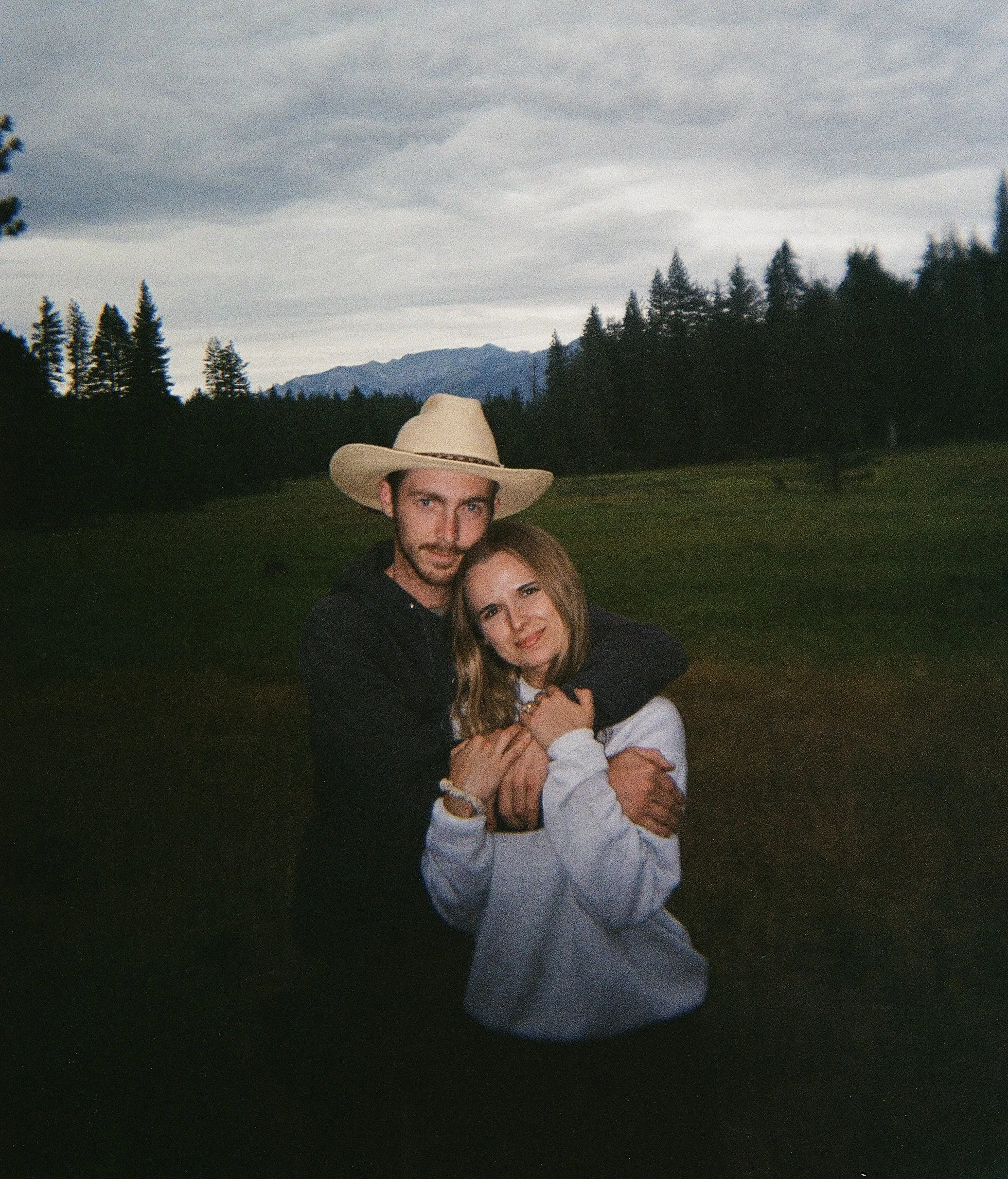 A couple hugging outdoors, with forest and mountain landscape in the background. The man is wearing a straw hat and a black jacket, and the woman has light brown hair and is wearing a white top.