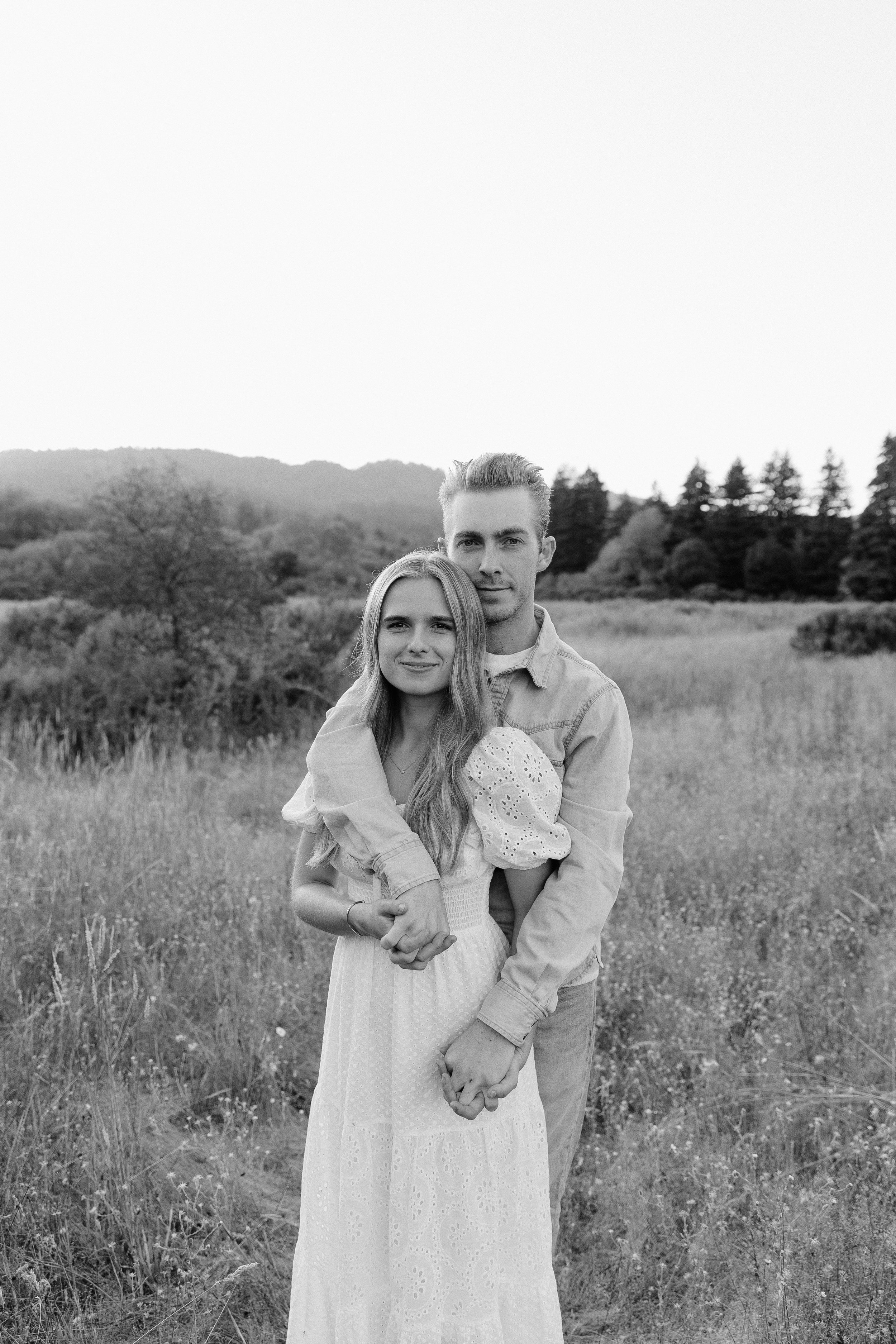 A black and white photo of a young couple standing in a grassy field with trees and mountains in the background. They are embracing, with the woman smiling and the man looking at the camera.
