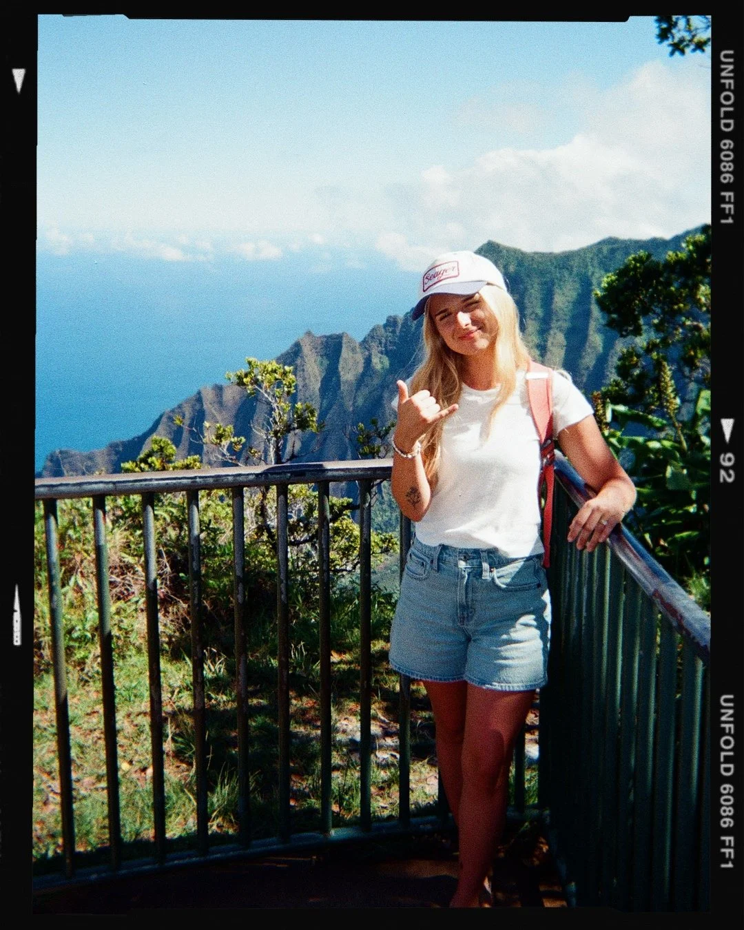 A woman with blonde hair wearing a white t-shirt, denim shorts, a white cap, and a pink backpack, standing by a railing with a mountainous landscape and the ocean behind her.