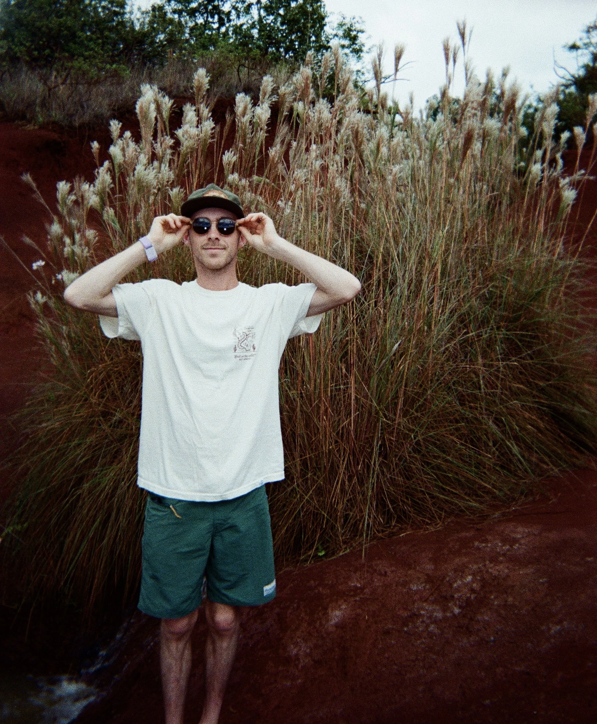 A young man wearing sunglasses, a white t-shirt, and dark shorts stands in front of tall, dry grasses with trees in the background. He is adjusting his sunglasses with both hands.