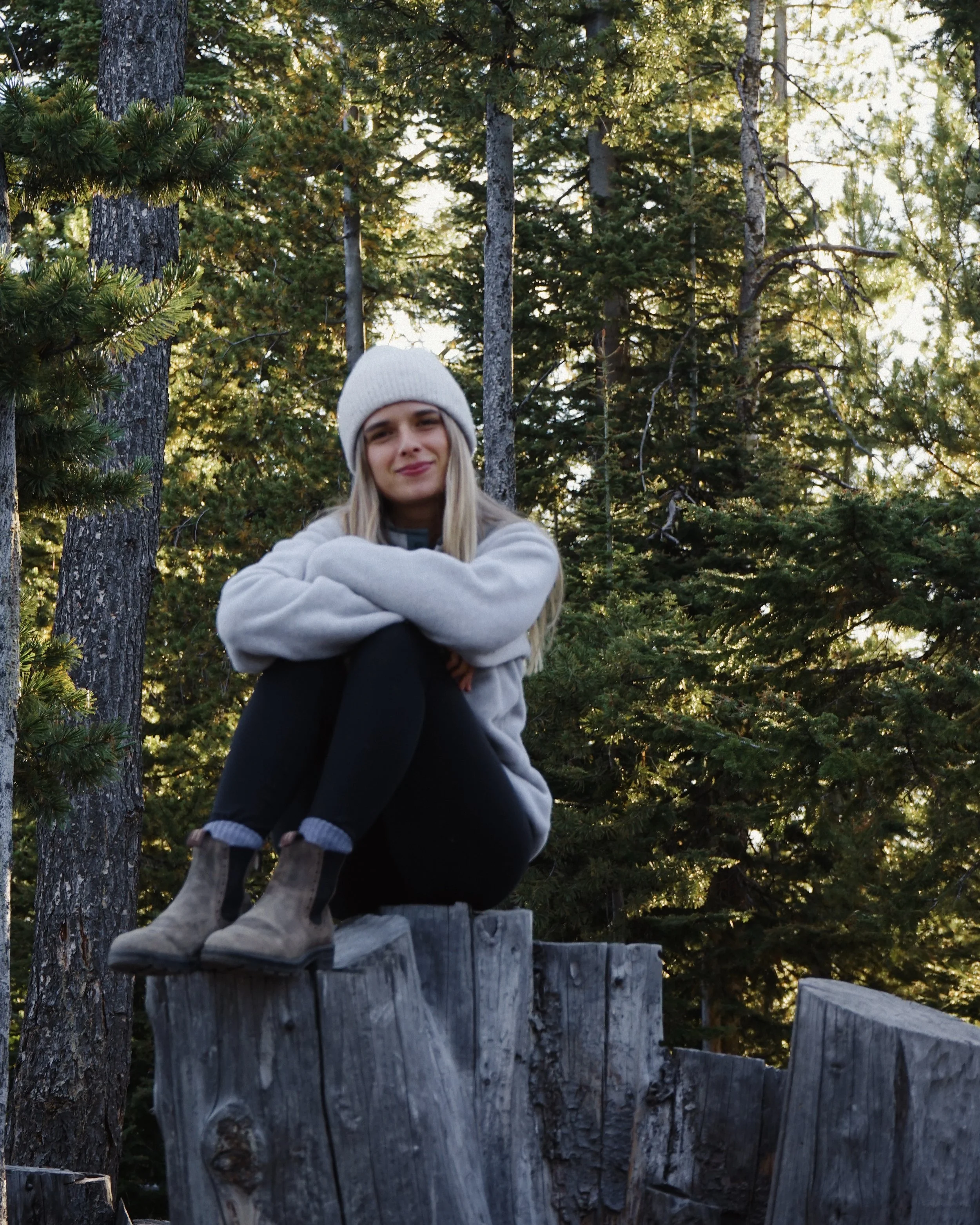 A young woman sitting on a wooden platform in forest surrounded by tall trees and green foliage, wearing a white beanie, a light gray sweatshirt, and black pants, with boots, looking at the camera with a slight smile.