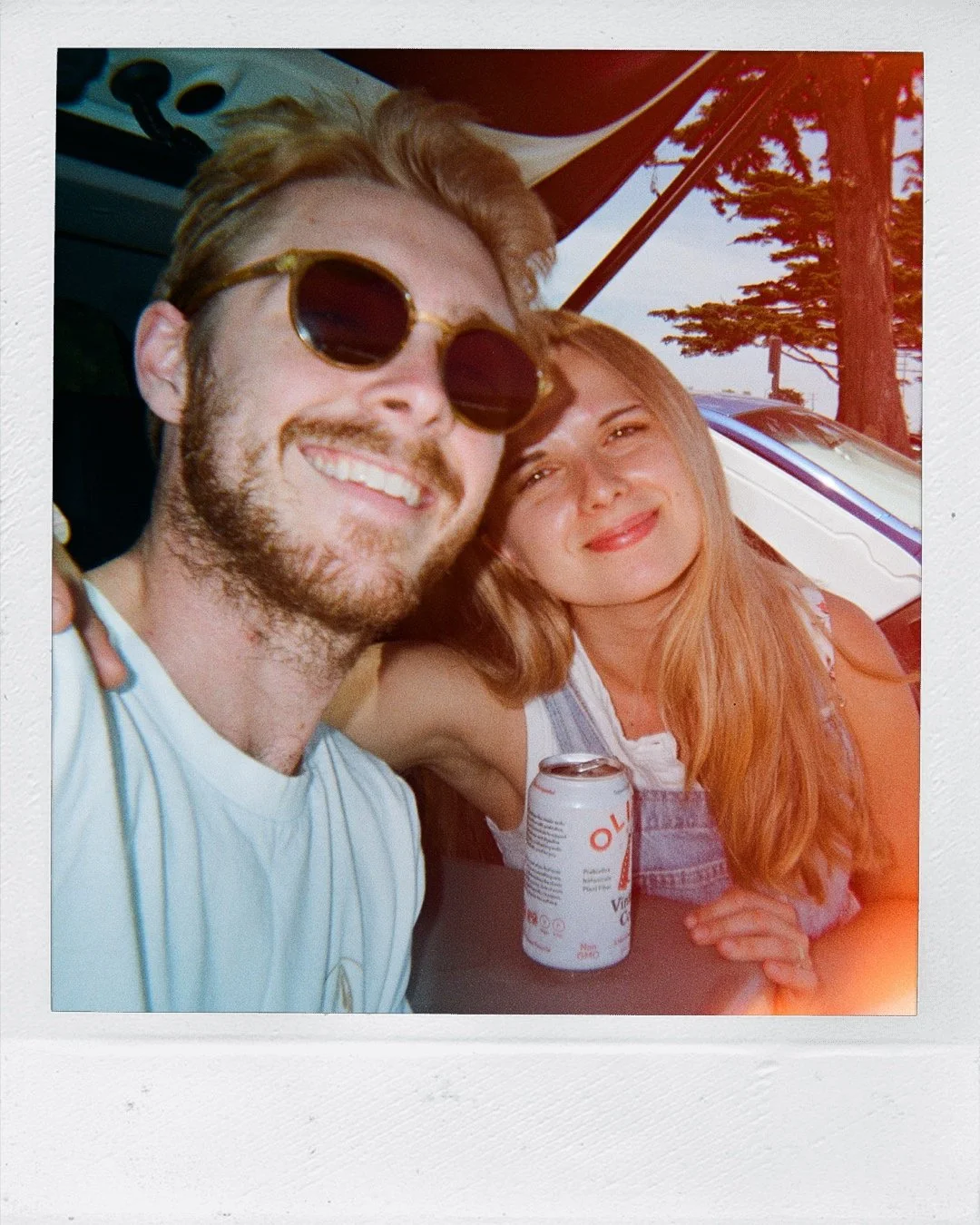 A man with blonde hair, sunglasses, and a beard smiling next to a woman with long red hair at an outdoor location with trees and a car in the background, holding a soda can.