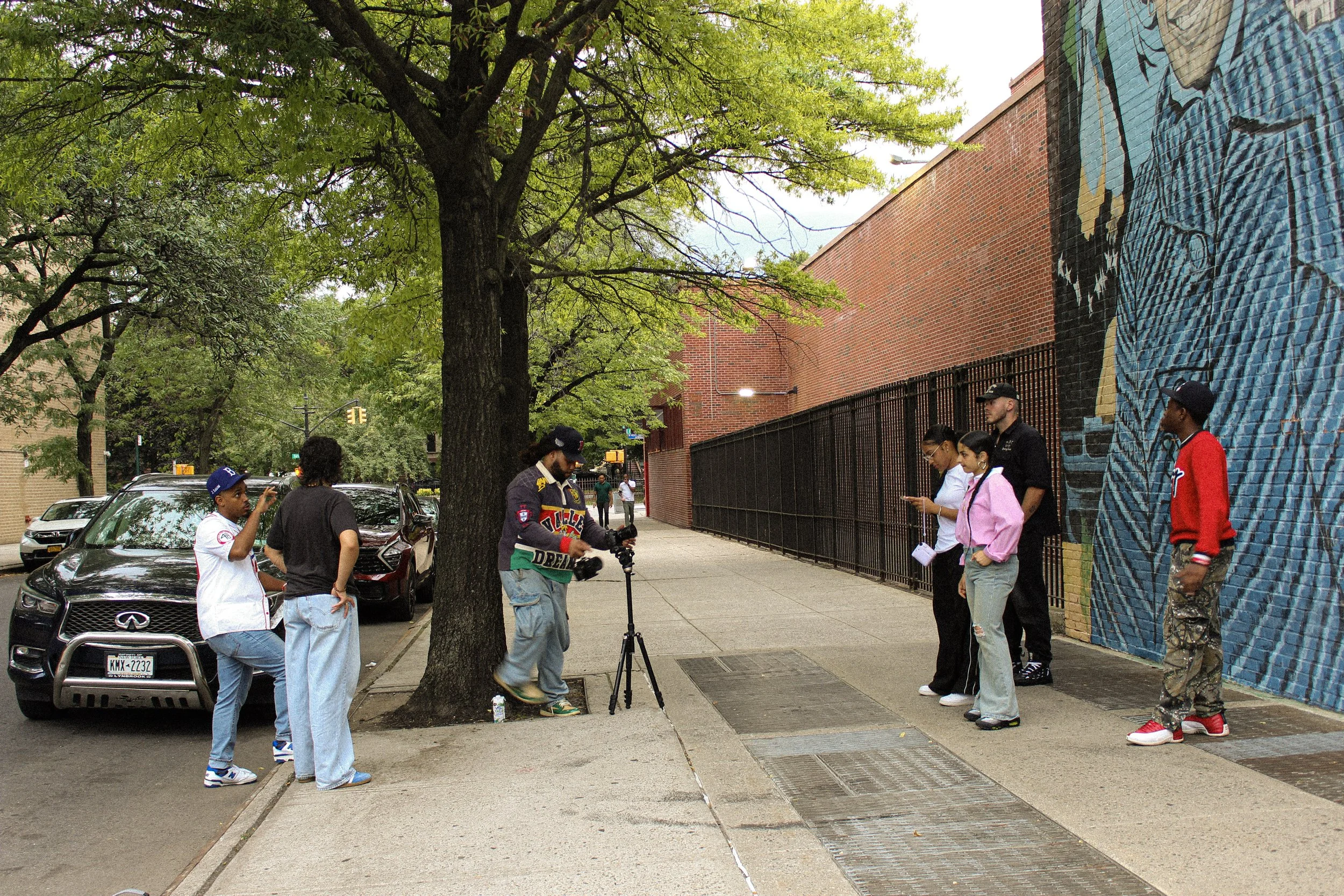 A group of people in urban street scene standing along sidewalk near a large tree and colorful mural on brick wall.