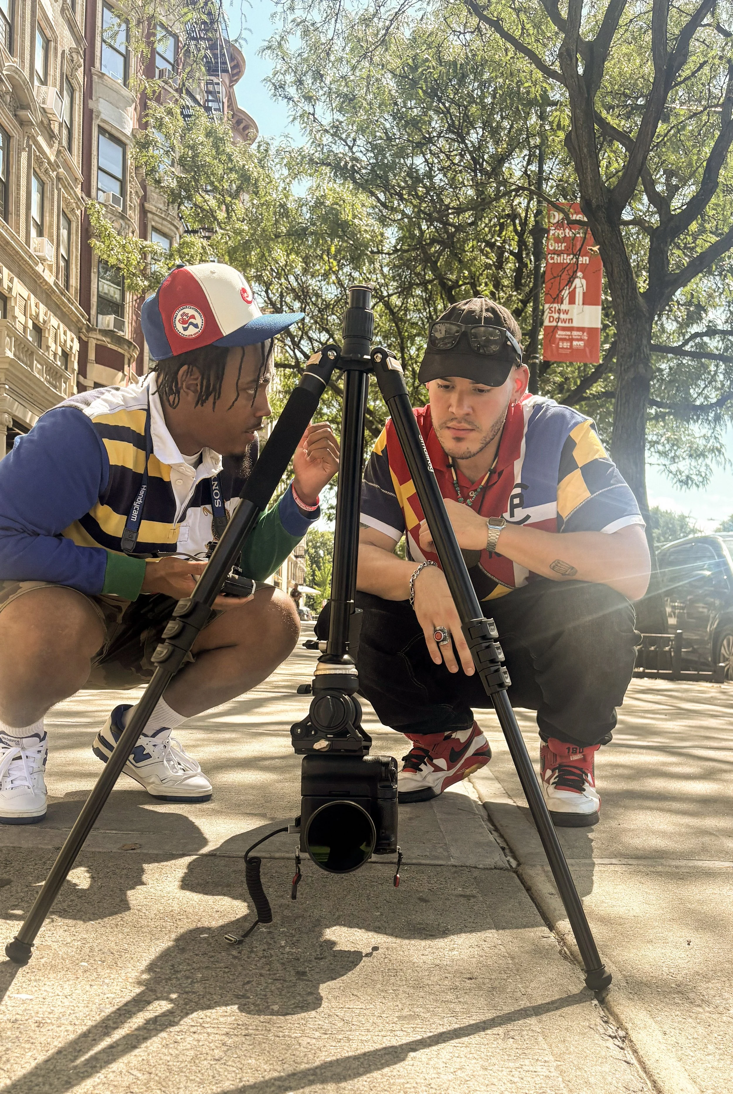 Two young men are working with a camera mounted on a tripod on a city sidewalk. They are crouched down, focusing intently on the camera, with a backdrop of trees, historic buildings, and a sunny sky.