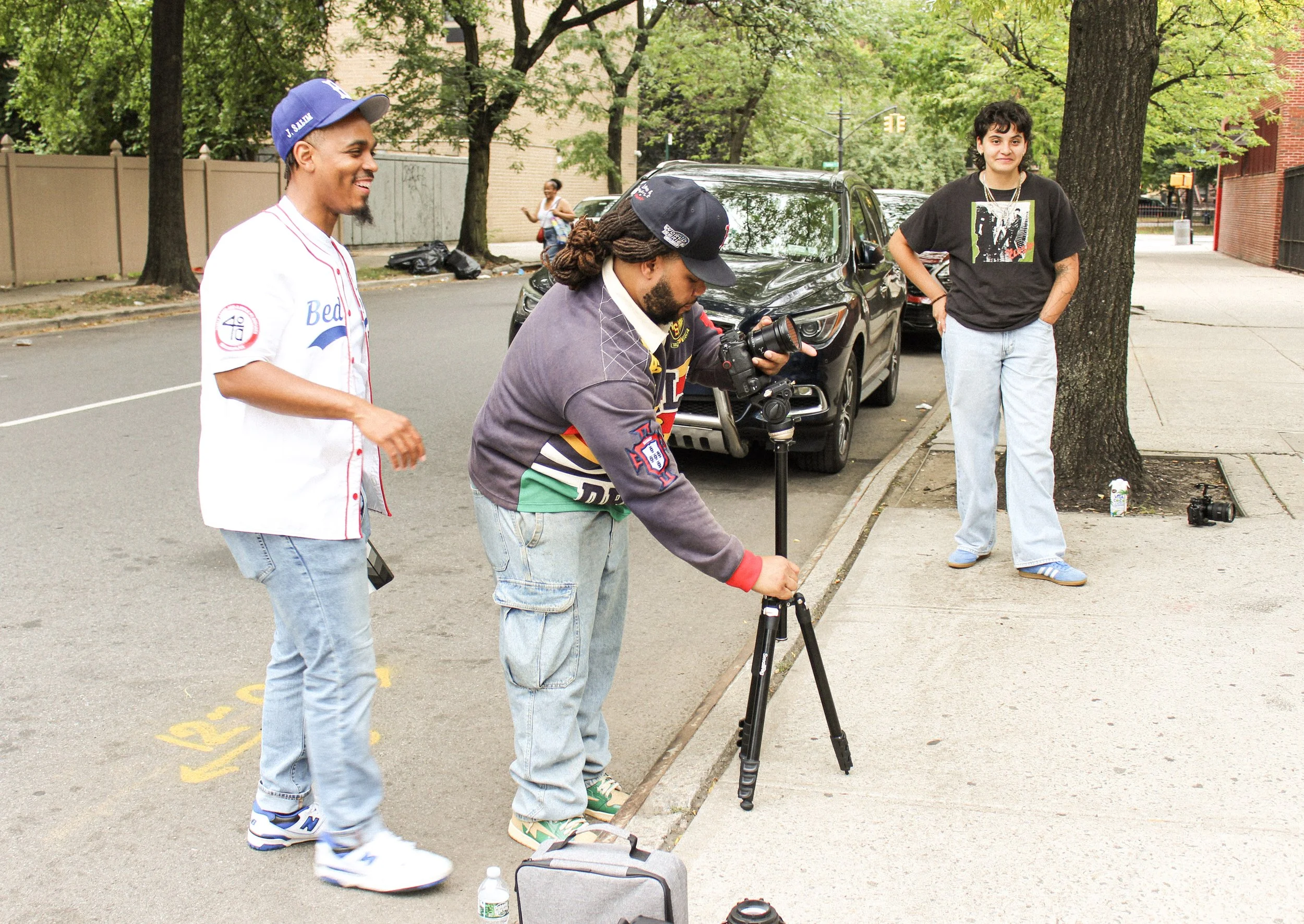 Three young people are standing on a city sidewalk near a parked black car and a tree, with one person adjusting camera equipment, while another is holding a camera and the third is leaning against the tree.