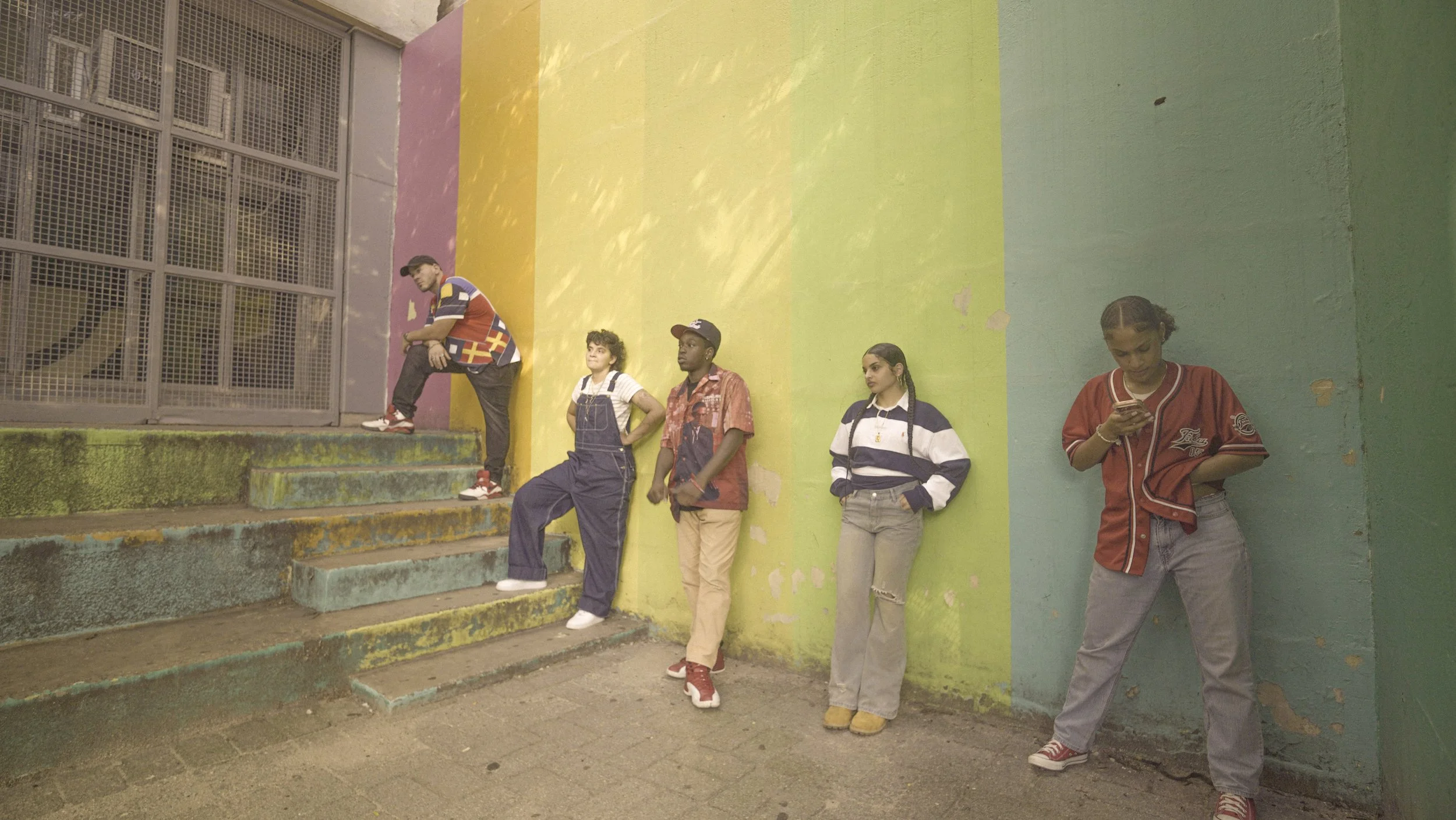 Five young people are standing against a colorful, striped wall by a set of worn metal steps. The wall has peeling paint in pink, yellow, and green. The group is in casual clothing, with some looking at their phones and others looking away.
