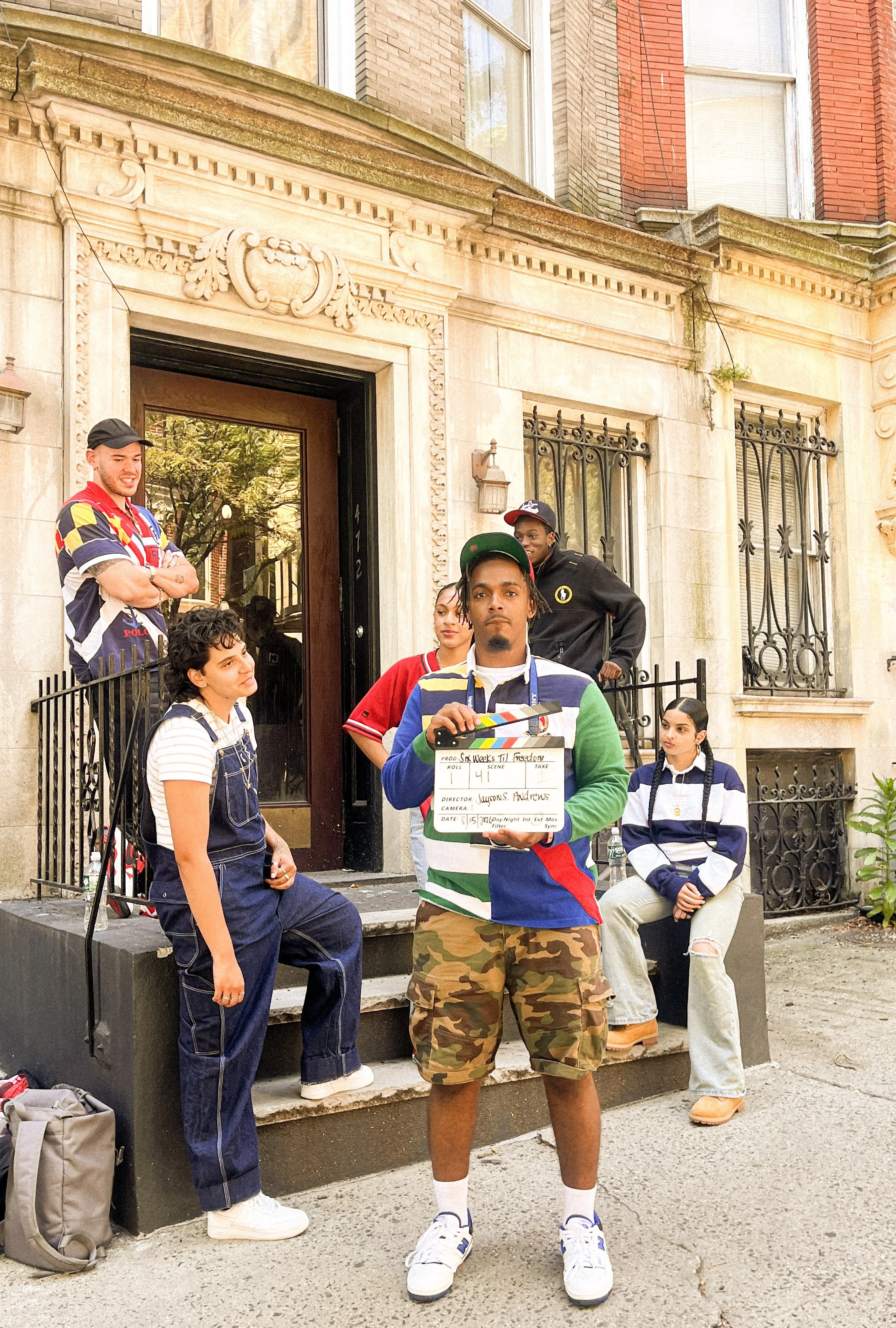 Group of diverse young people standing and sitting outside a brownstone building with ornate architectural details, some wearing casual clothes and jackets, one person holding a clapperboard, appearing to be on a film set.
