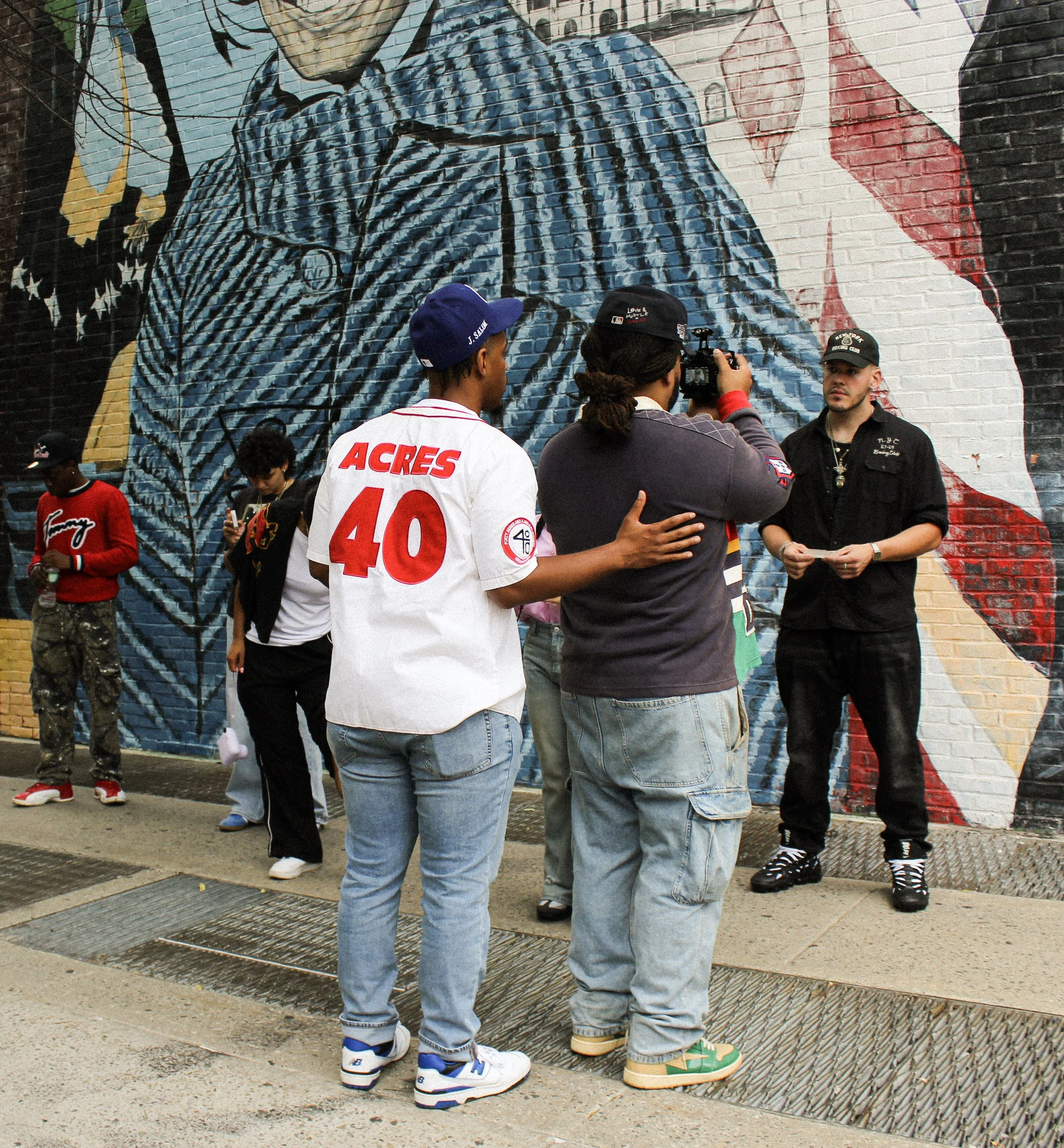 Group of young people gathered in front of a large mural on a brick wall, some taking photos or looking at each other. The mural depicts a person's face with glasses, and a red and white background. The people are dressed casually, with some wearing 