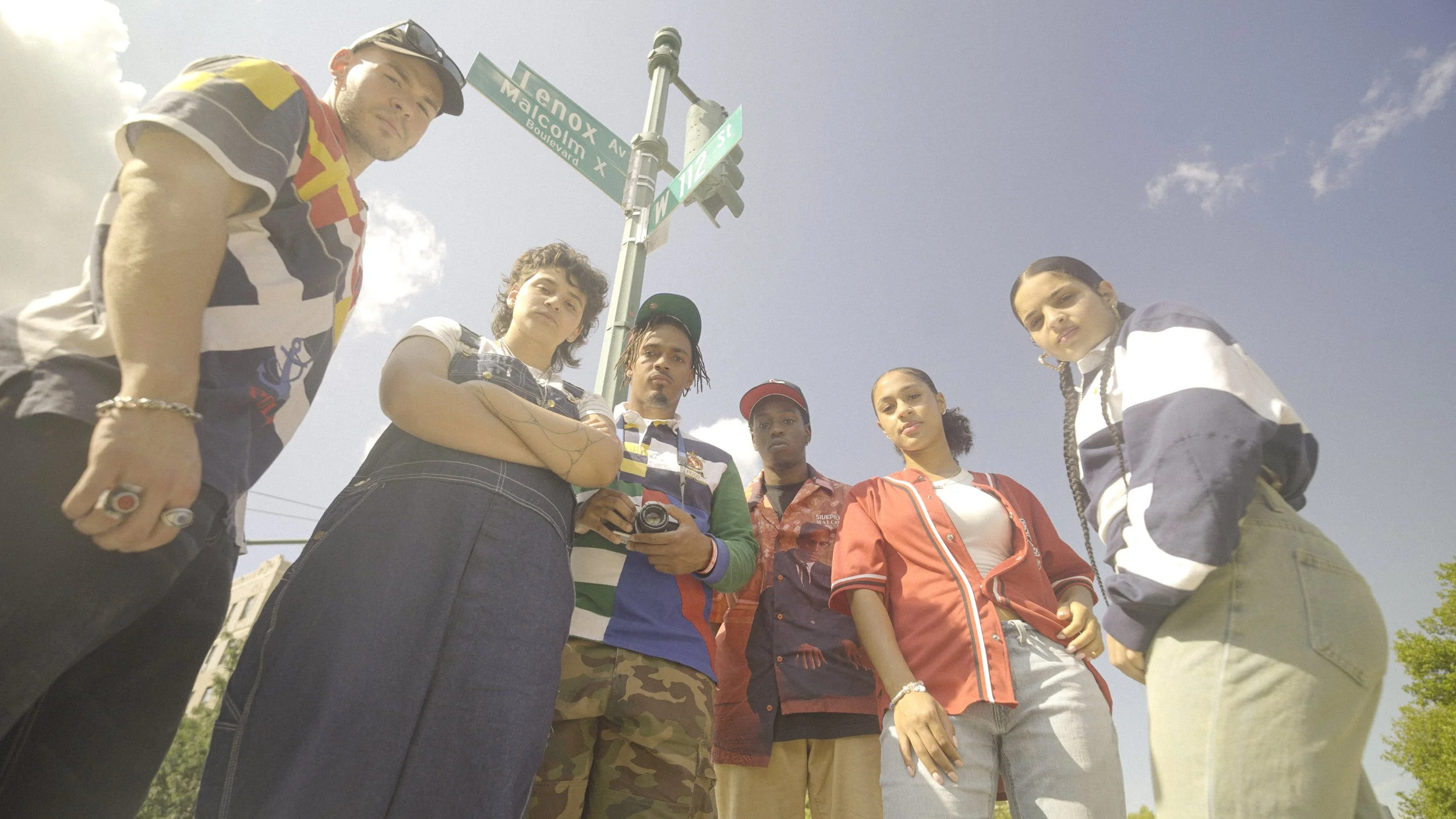 Group of six young adults standing together outdoors under street signs at the intersection of Menox Avenue and Malcolm X Boulevard, looking down at the camera.