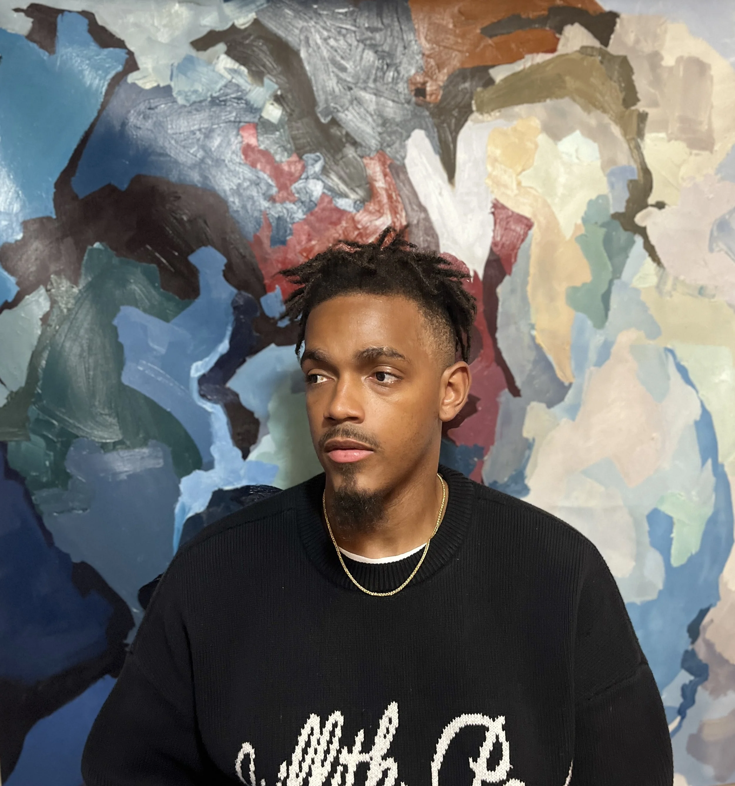 A young man with dark hair and a light beard is looking slightly to his left. He is wearing a black sweater, a gold chain around his neck, and a black and white shirt with some cursive writing. The background is a colorful abstract mural with shades of blue, gray, white, black, and some reddish hues.