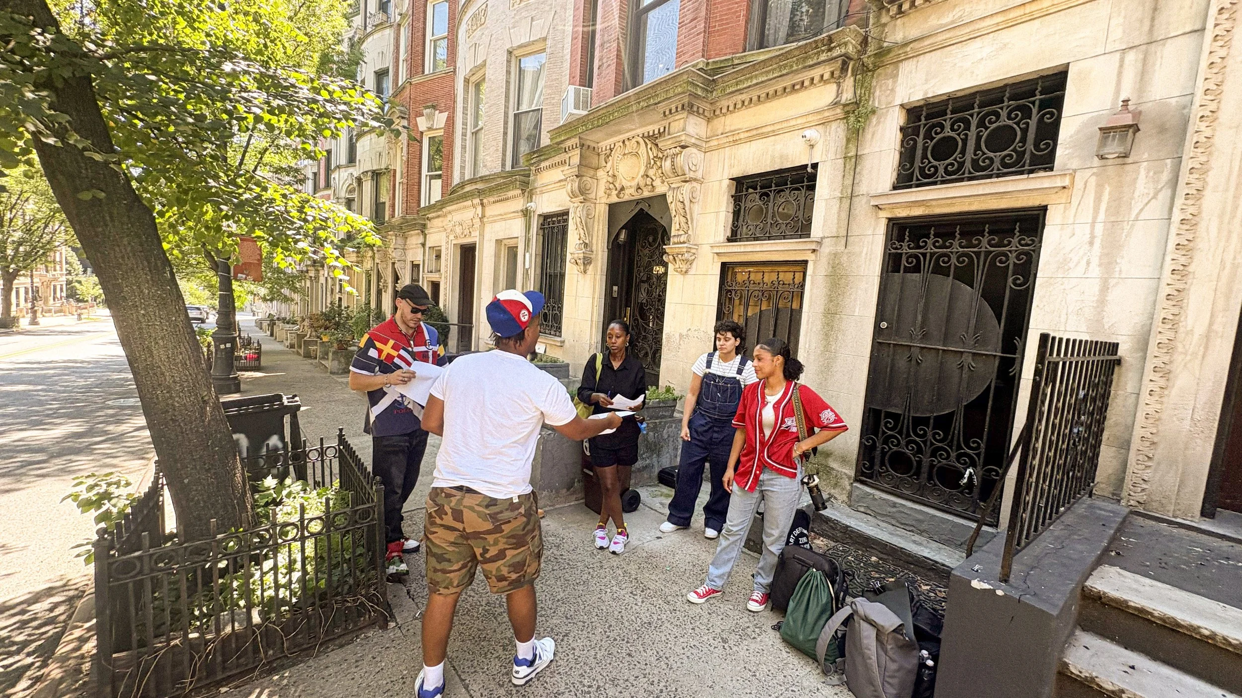A group of five people standing outside on a city sidewalk in front of an ornate building entrance, engaged in conversation, with trees lining the street and black wrought iron fencing.