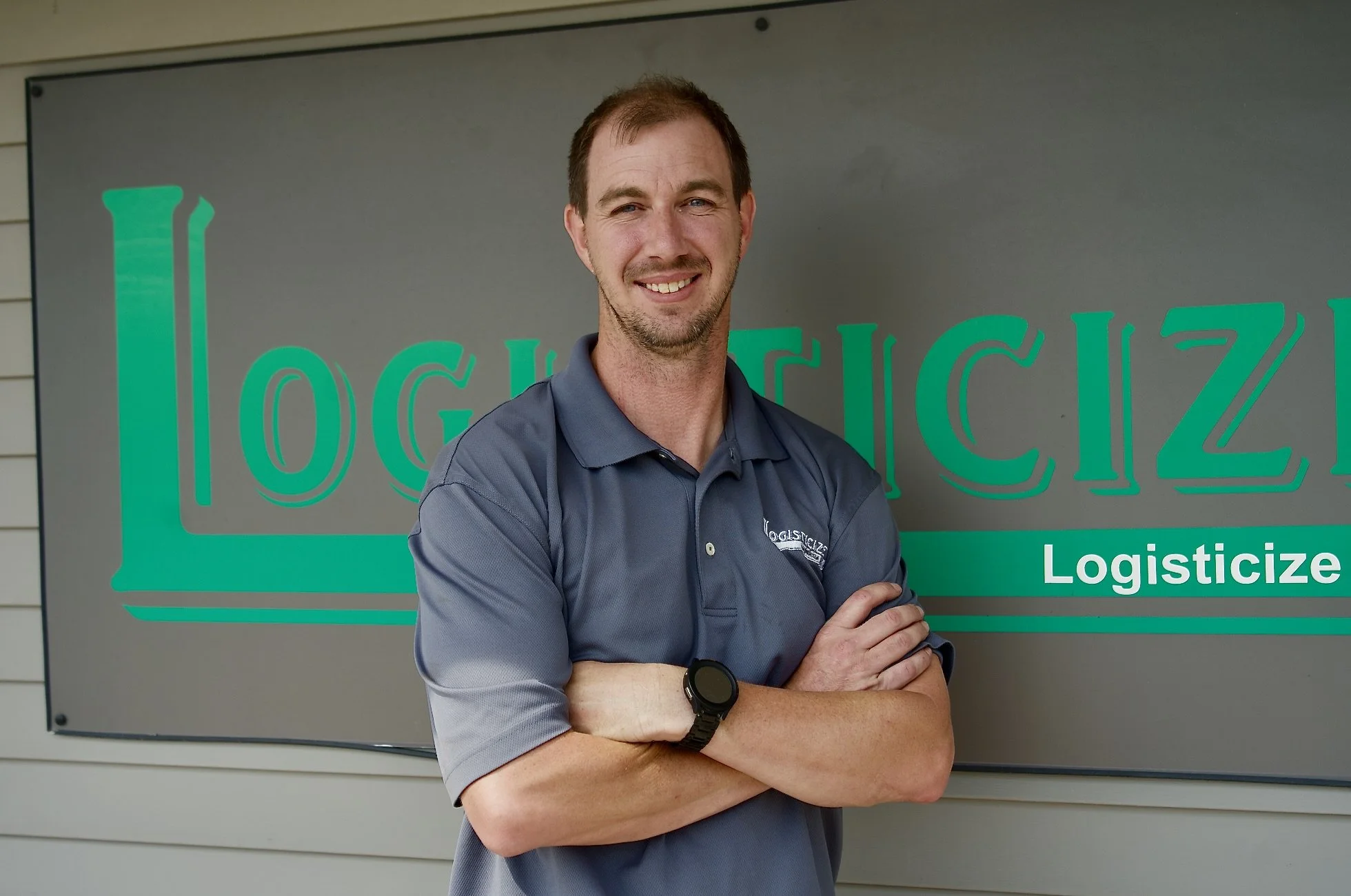 A man with short brown hair and a beard, smiling and standing with arms crossed in front of a green and gray sign that says "Logisticize." He is wearing a gray collared shirt and a black smartwatch on his left wrist.