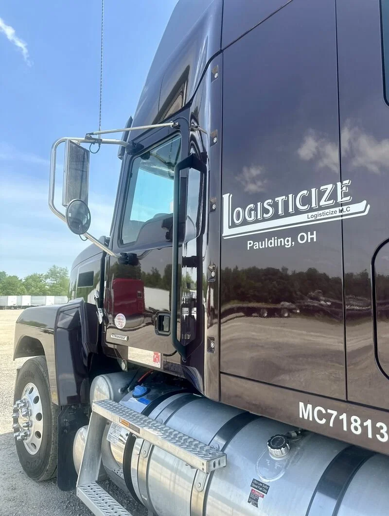 Close-up of a black logistics truck with the text 'Logisticize, Paulding, Ohio' on the side, parked outdoors under a partly cloudy sky.