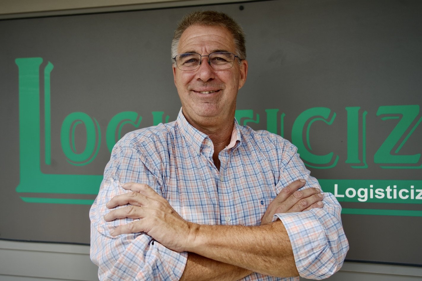 A middle-aged man wearing glasses and a plaid shirt standing with arms crossed in front of a sign that reads 'Logisticiz' with smaller text underneath.