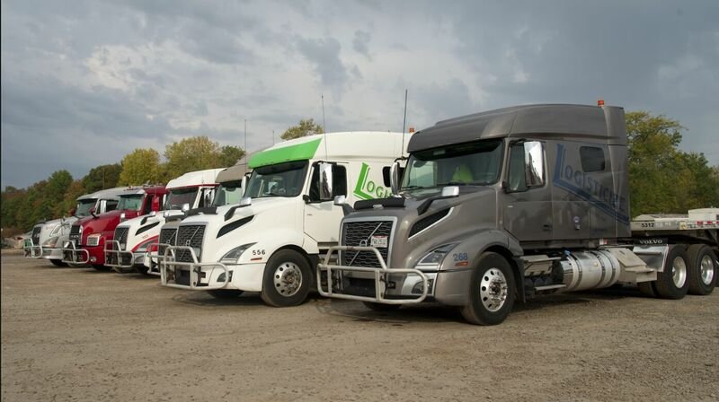 A lineup of semi-trucks parked on a dirt lot, with trees and cloudy sky in the background.