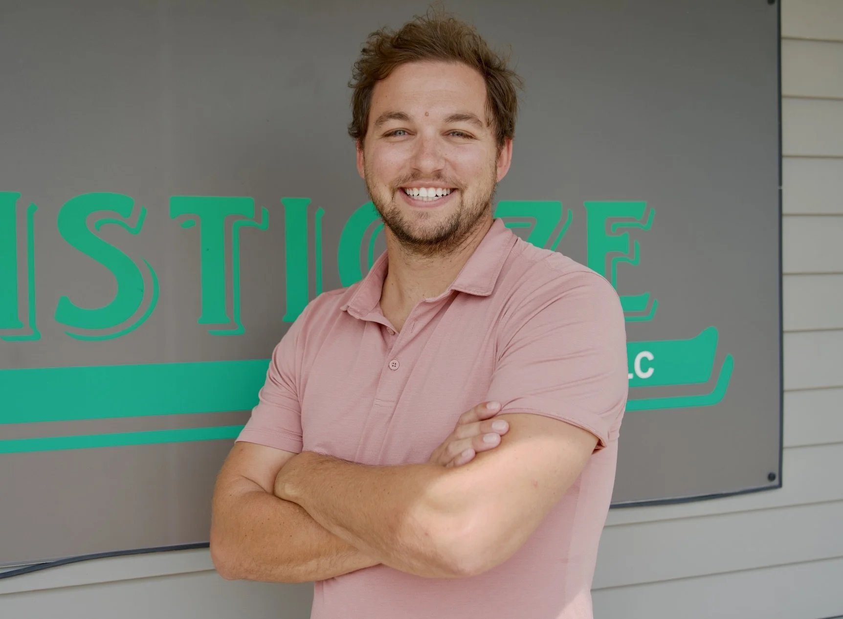 A smiling young man with light skin, brown curly hair, and a beard, wearing a pink polo shirt, standing in front of a sign that says 'ESTI' in large green letters.