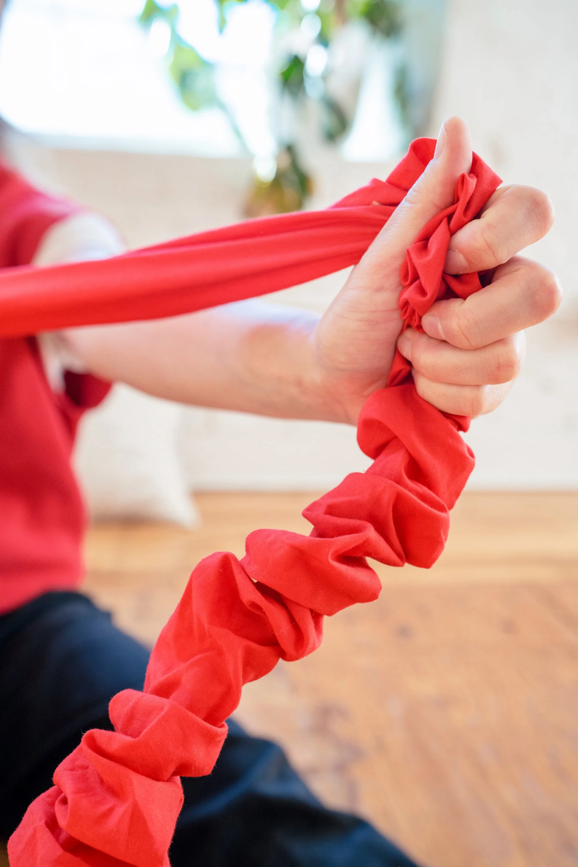 A person is twisting a red fabric or clothing with their hand. The focus is on the hand and fabric, with a blurred background of a room with plants and wooden flooring.