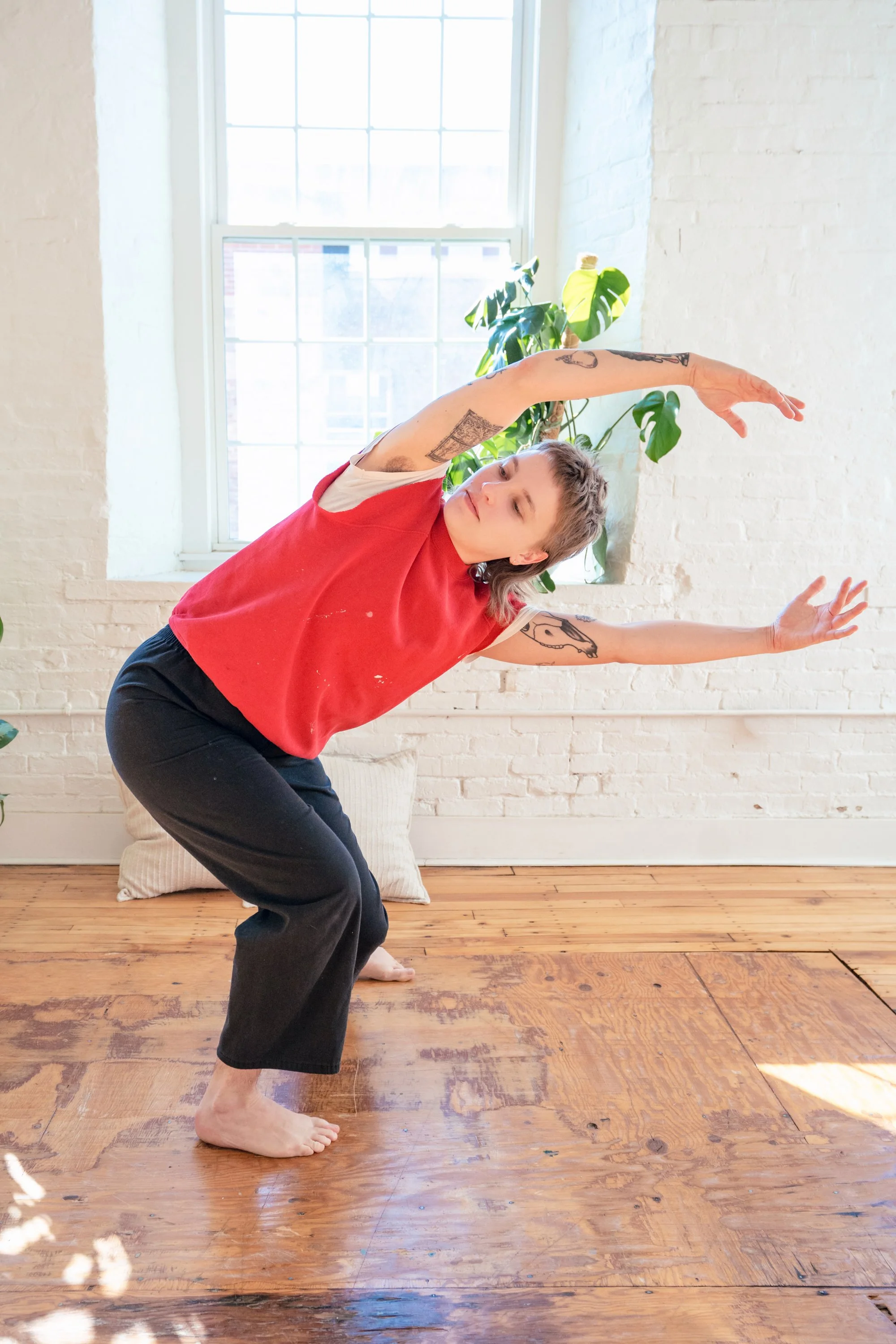 A person with tattoos dances in a bright room with white brick walls, a large window, hardwood floors, and a green plant.