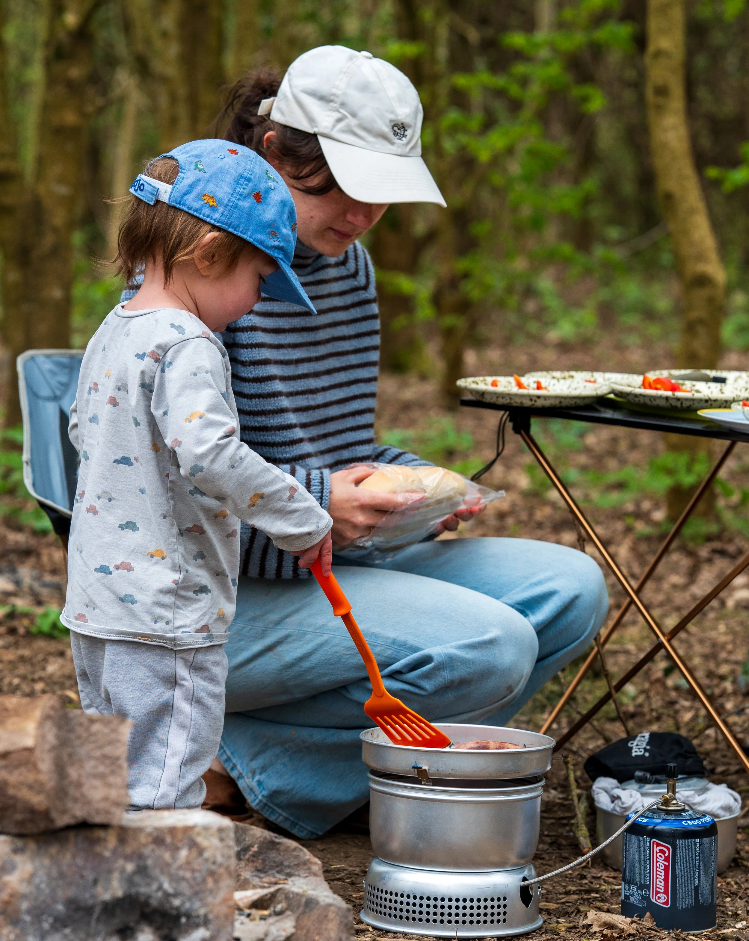 A woman and a child cooking outdoors in a forested area, with a camping stove and grilling supplies.