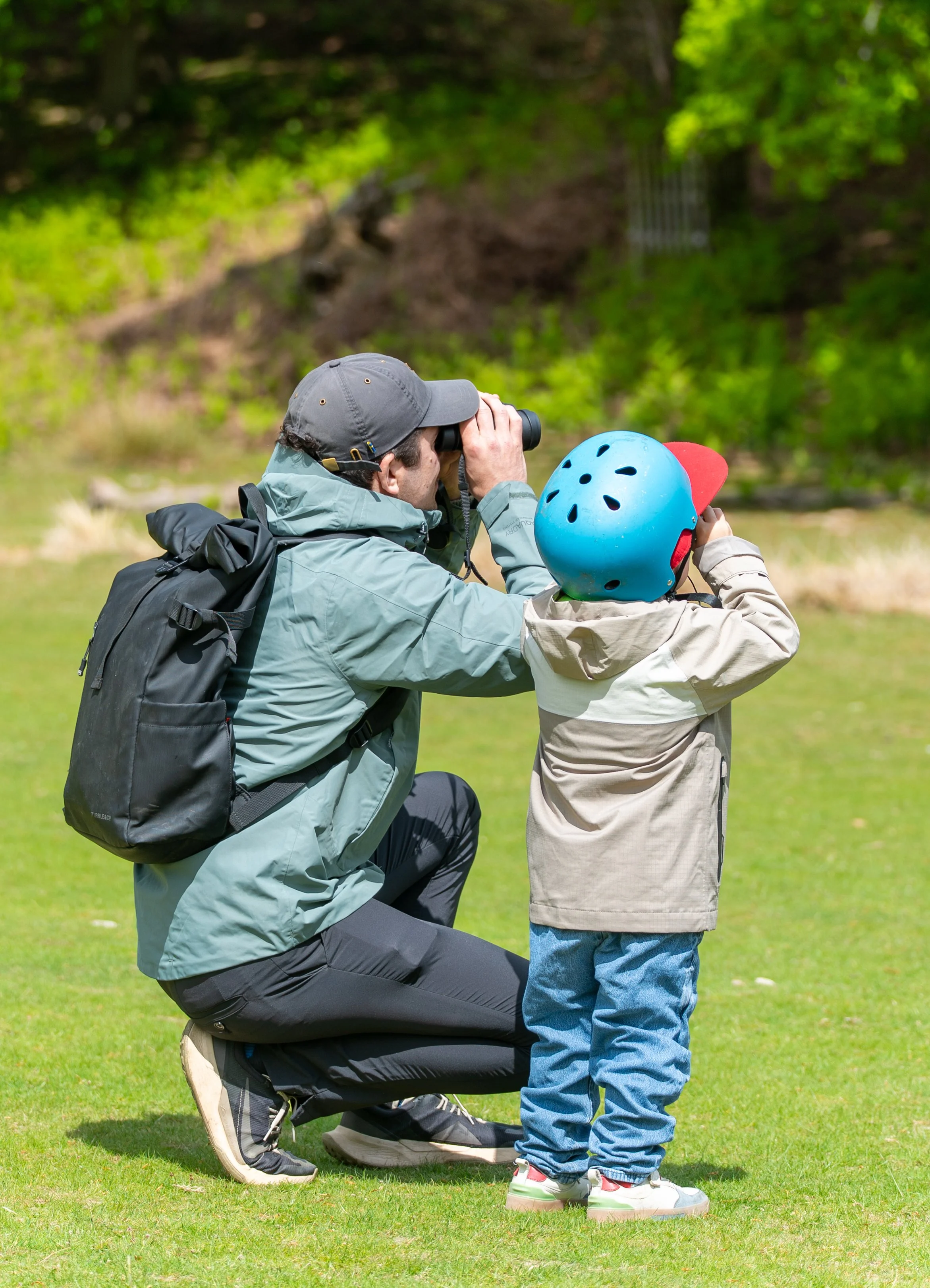 A man kneeling on one knee, holding a camera, taking a picture of a young boy wearing a blue helmet and a red cap, playing outdoors on a grassy field with trees in the background.