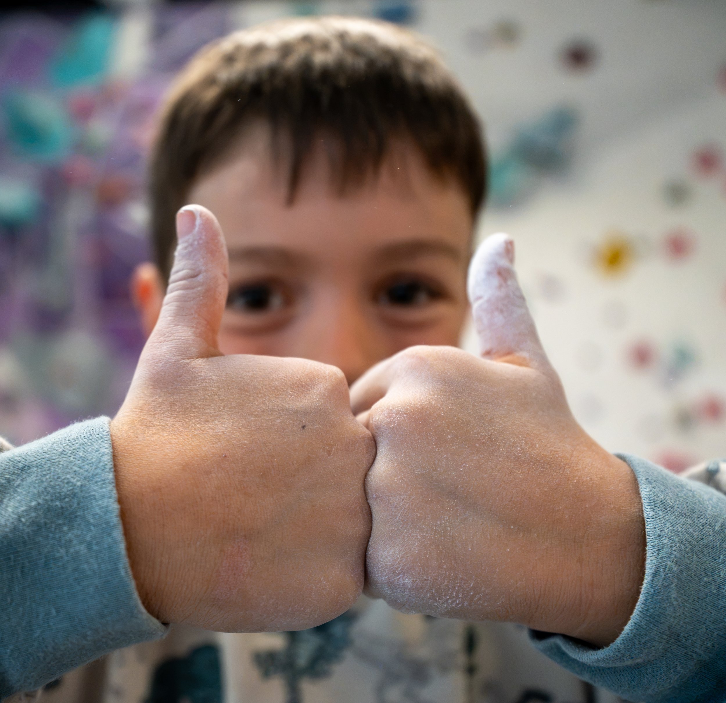 A child with short brown hair and dark eyes reaching out with fingers covered in white chalk, standing in front of a climbing wall with colorful holds.