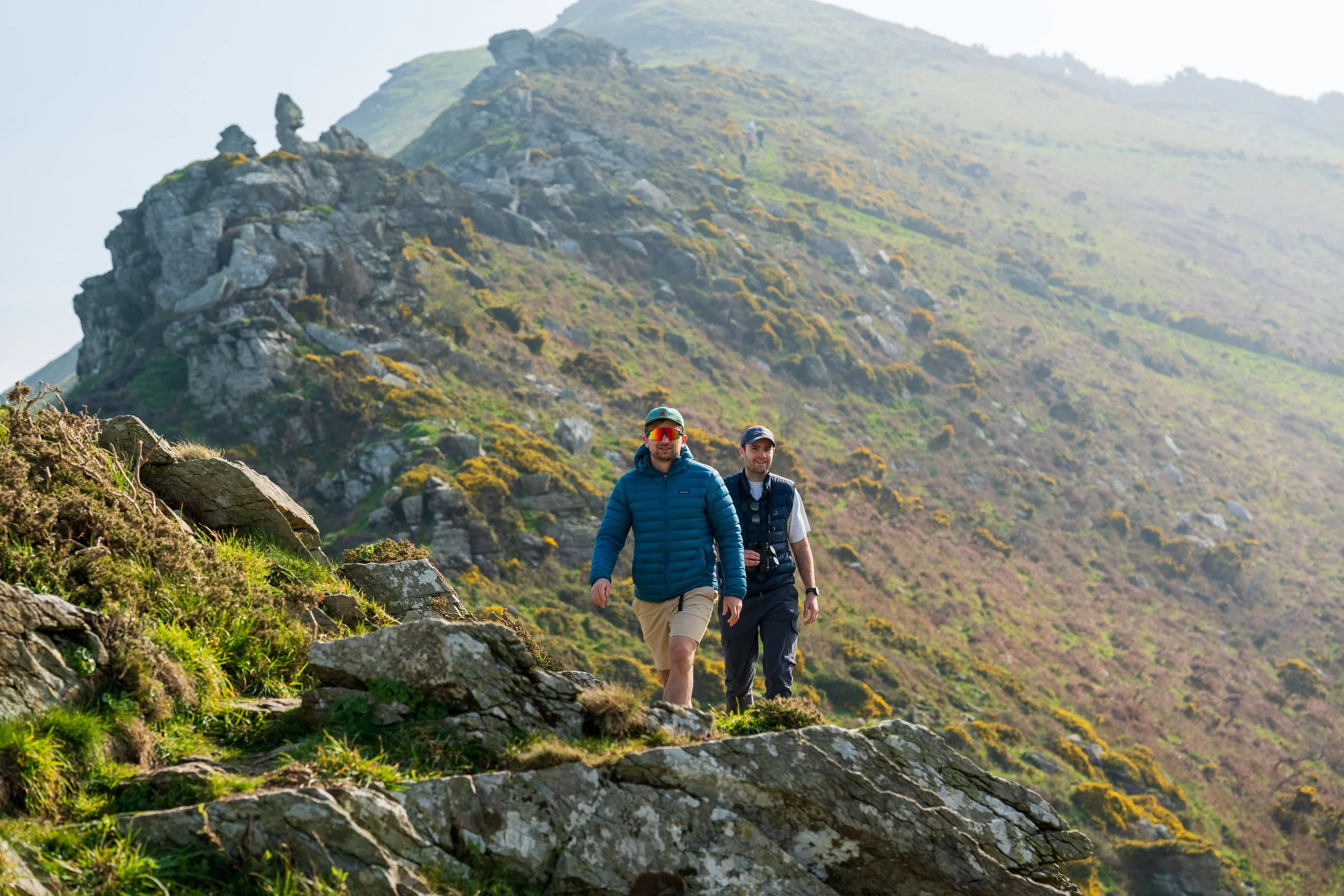Two men hiking on a trail through a green mountainous landscape with rocky terrain and hills in the background.