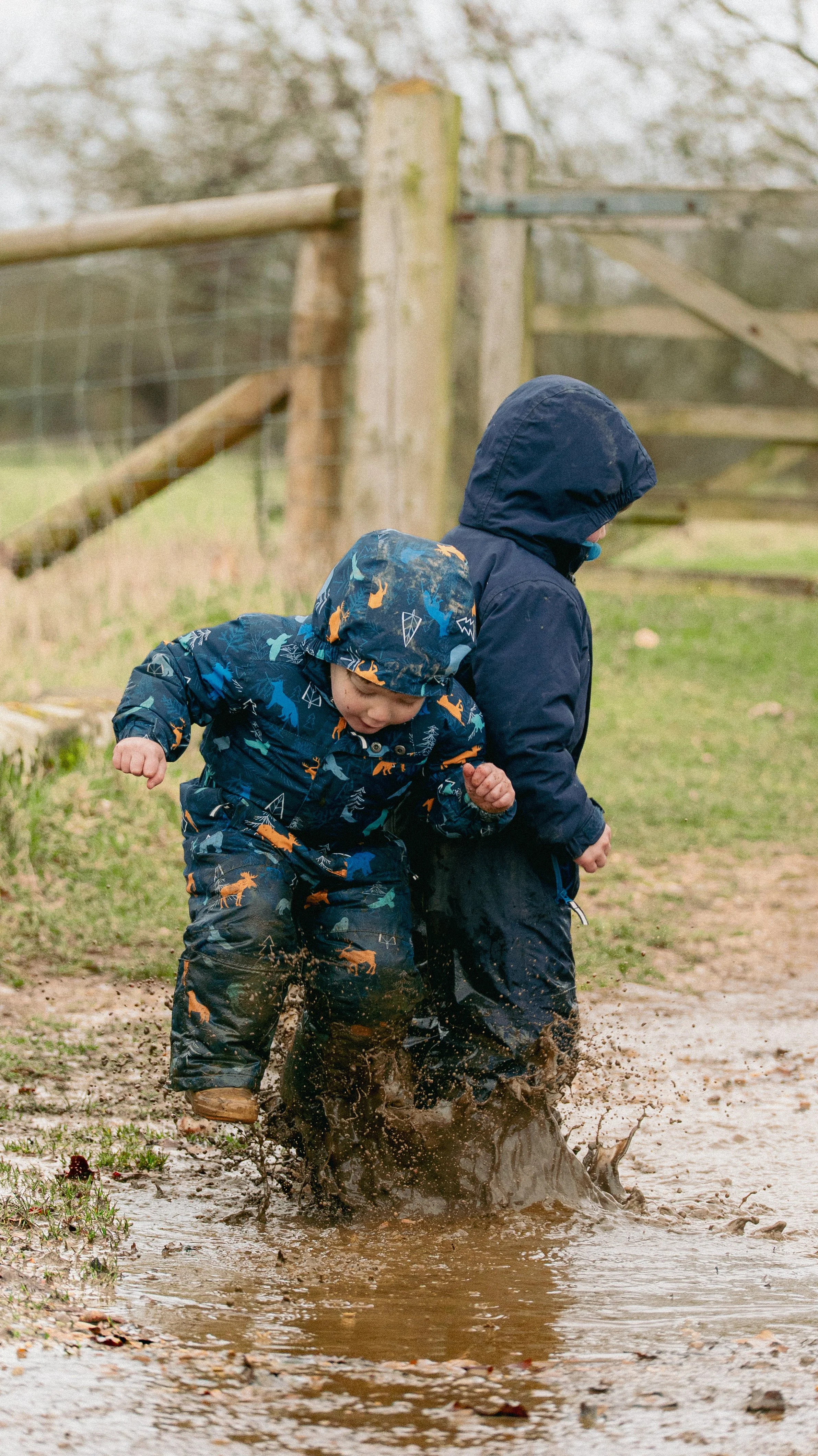 Two young children playing in a muddy puddle during rainy weather, dressed in waterproof jackets and pants, with one child's hood up, in an outdoor setting with a wooden fence in the background.