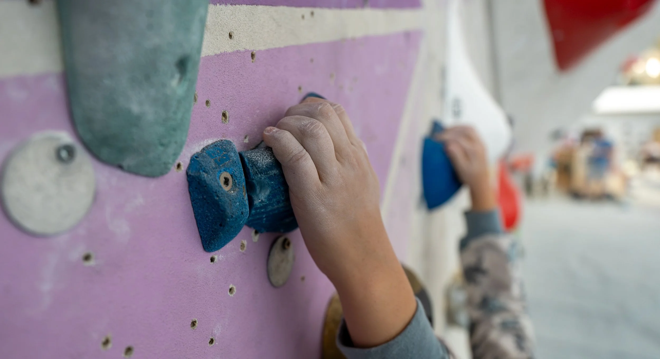 A person climbing an indoor bouldering wall using colorful holds, with other climbers visible in the background.