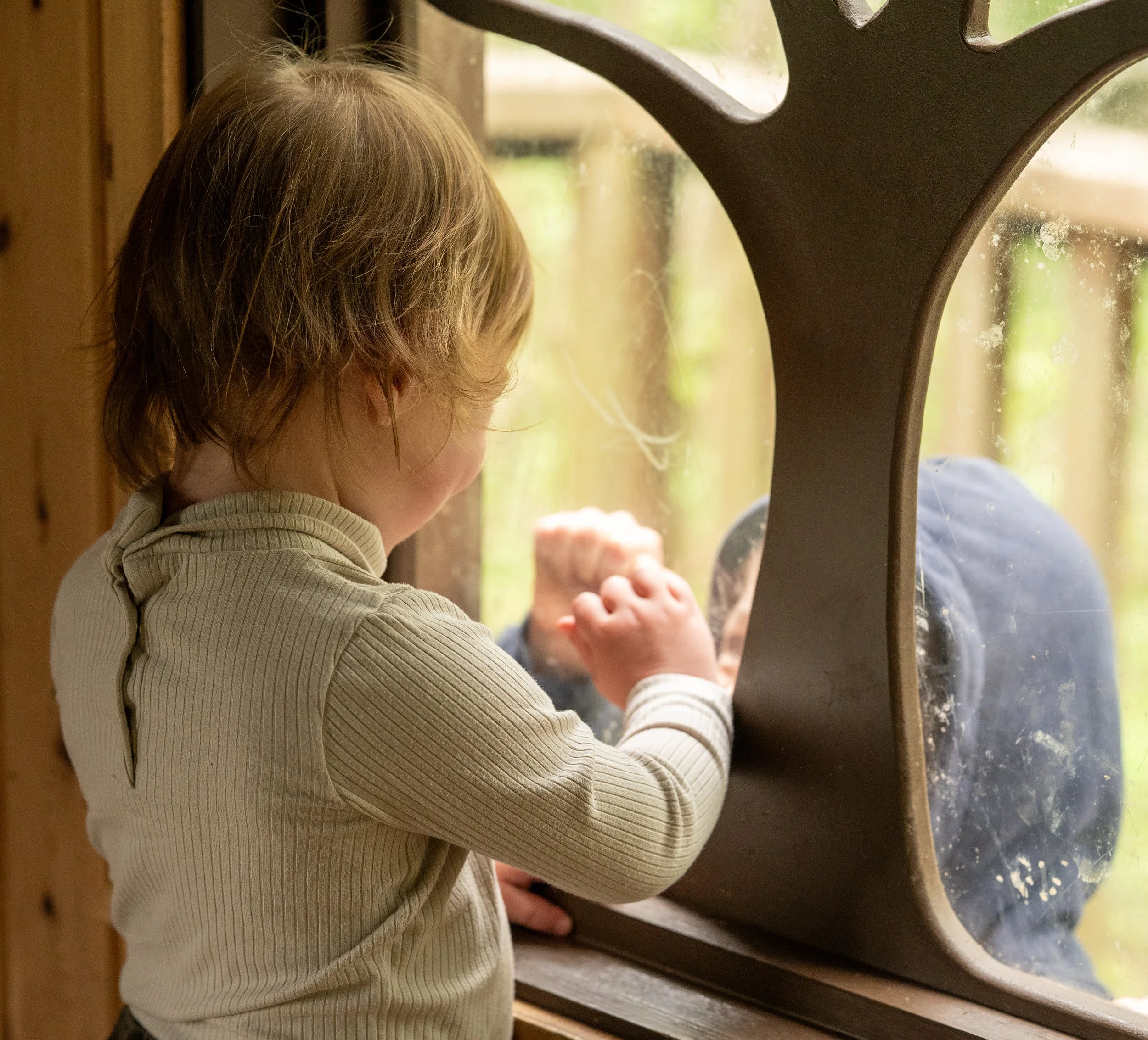 A young boy with light brown hair looking out a window with a curious expression, hand resting on the window sill.