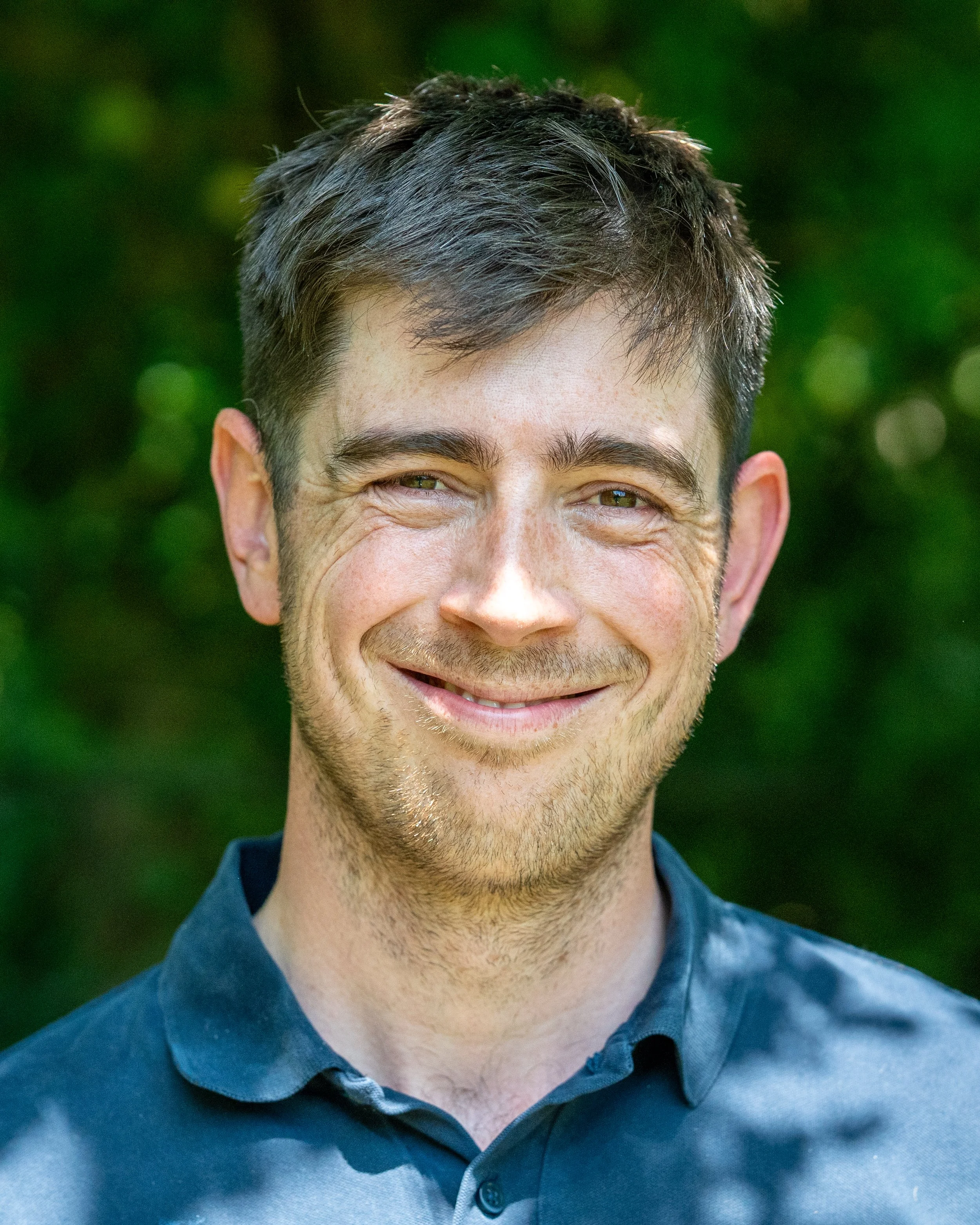 Close-up of a smiling man with short dark hair, light skin, and wearing a dark button-up shirt, outdoors with greenery in the background.