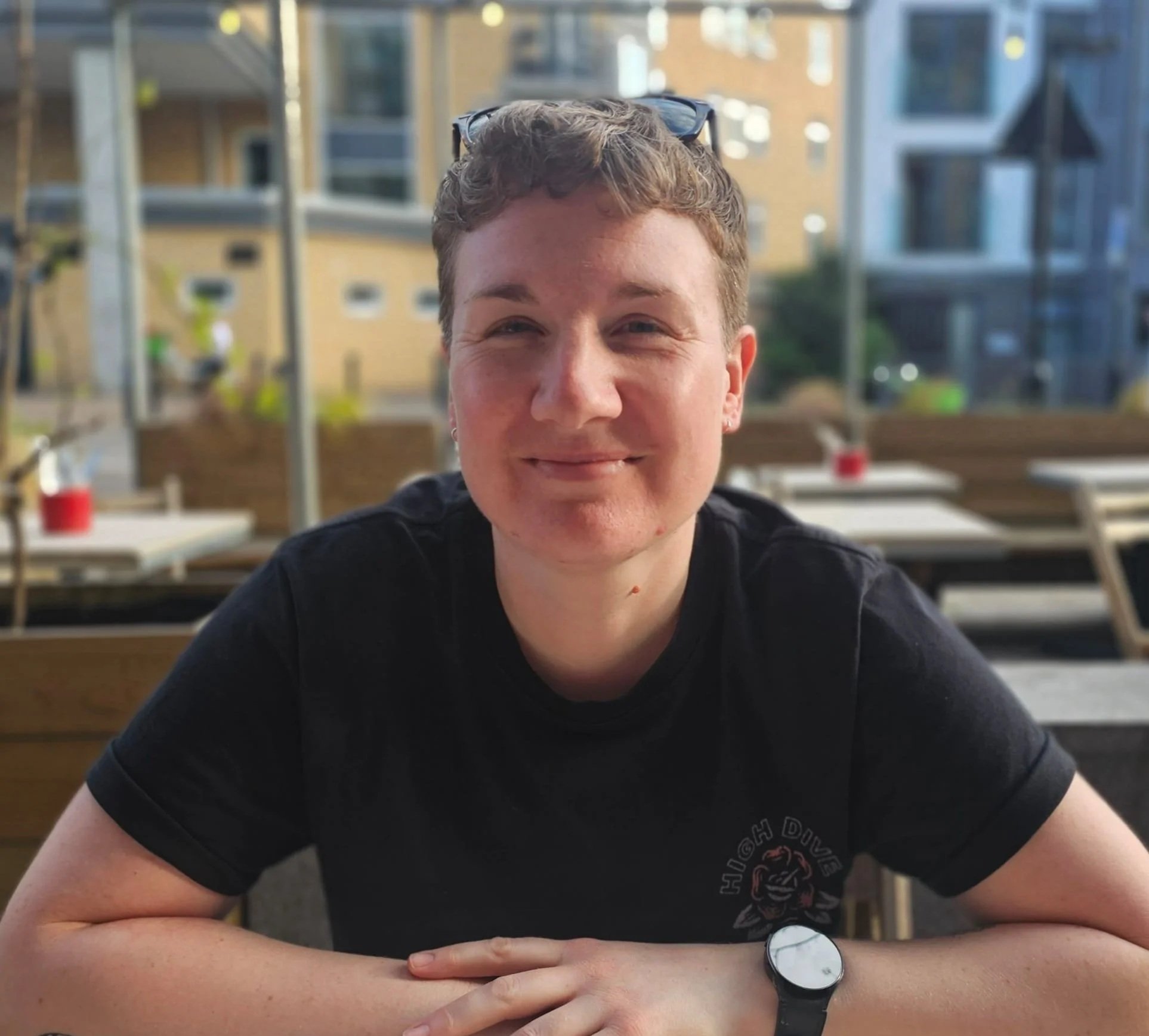 Close-up of a smiling young man with short curly hair wearing glasses on his head, sitting at an outdoor dining area with buildings in the background.