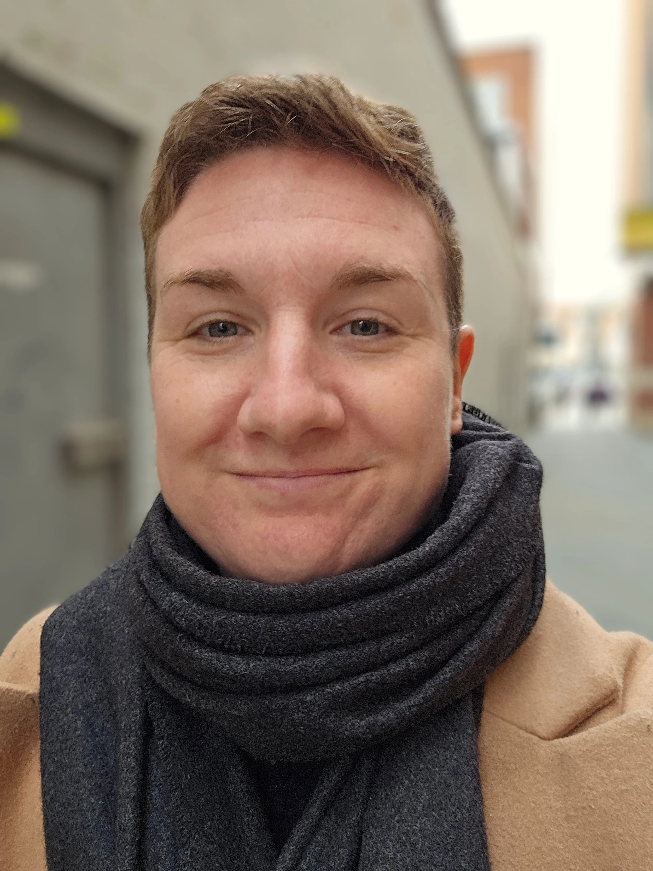 Close-up of a smiling person with short brown hair, wearing a dark scarf and a brown coat, indoor background.