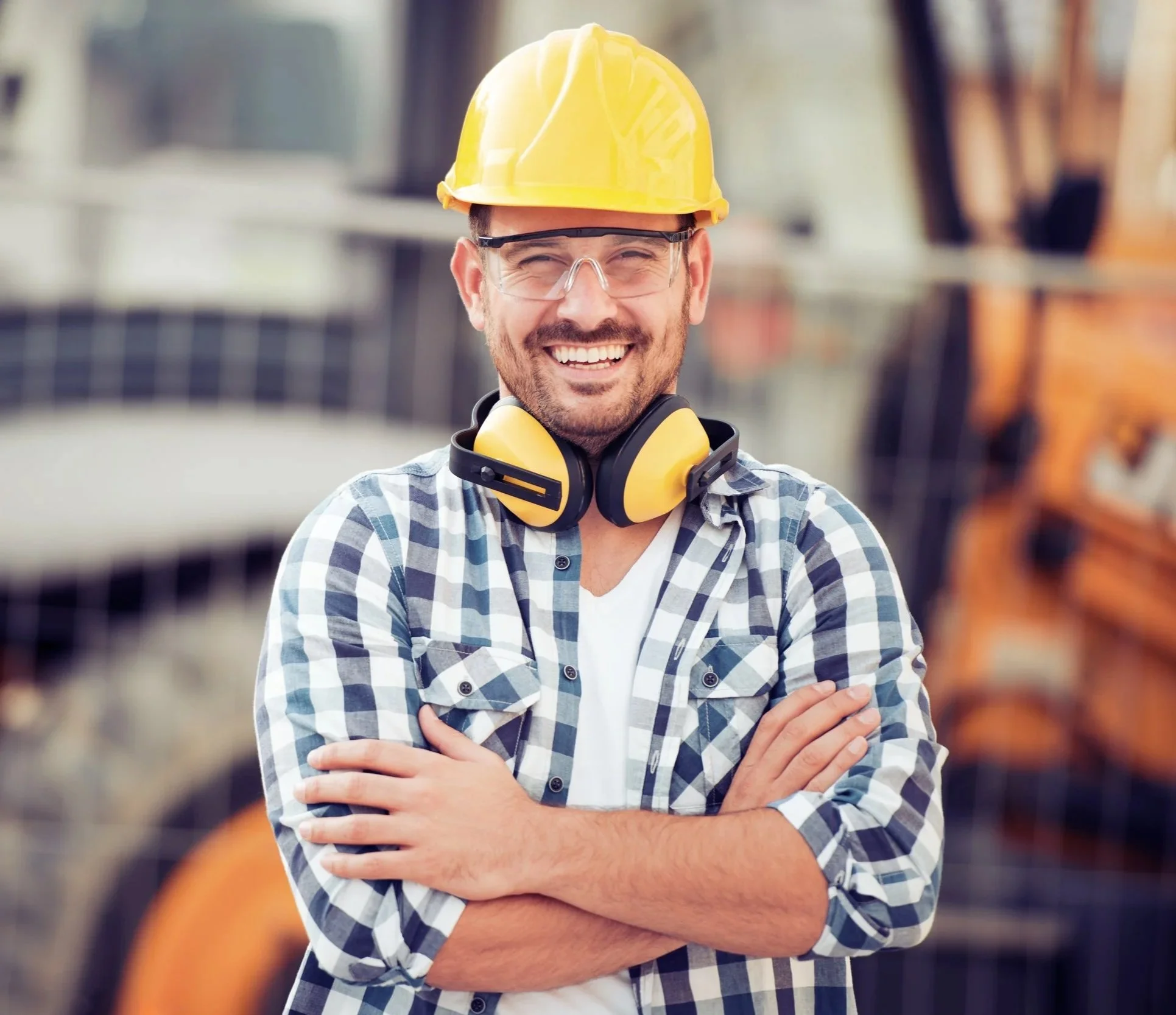 A cheerful man wearing a yellow construction helmet, safety glasses, and yellow ear protection around his neck, standing with arms crossed in a workshop or construction setting.