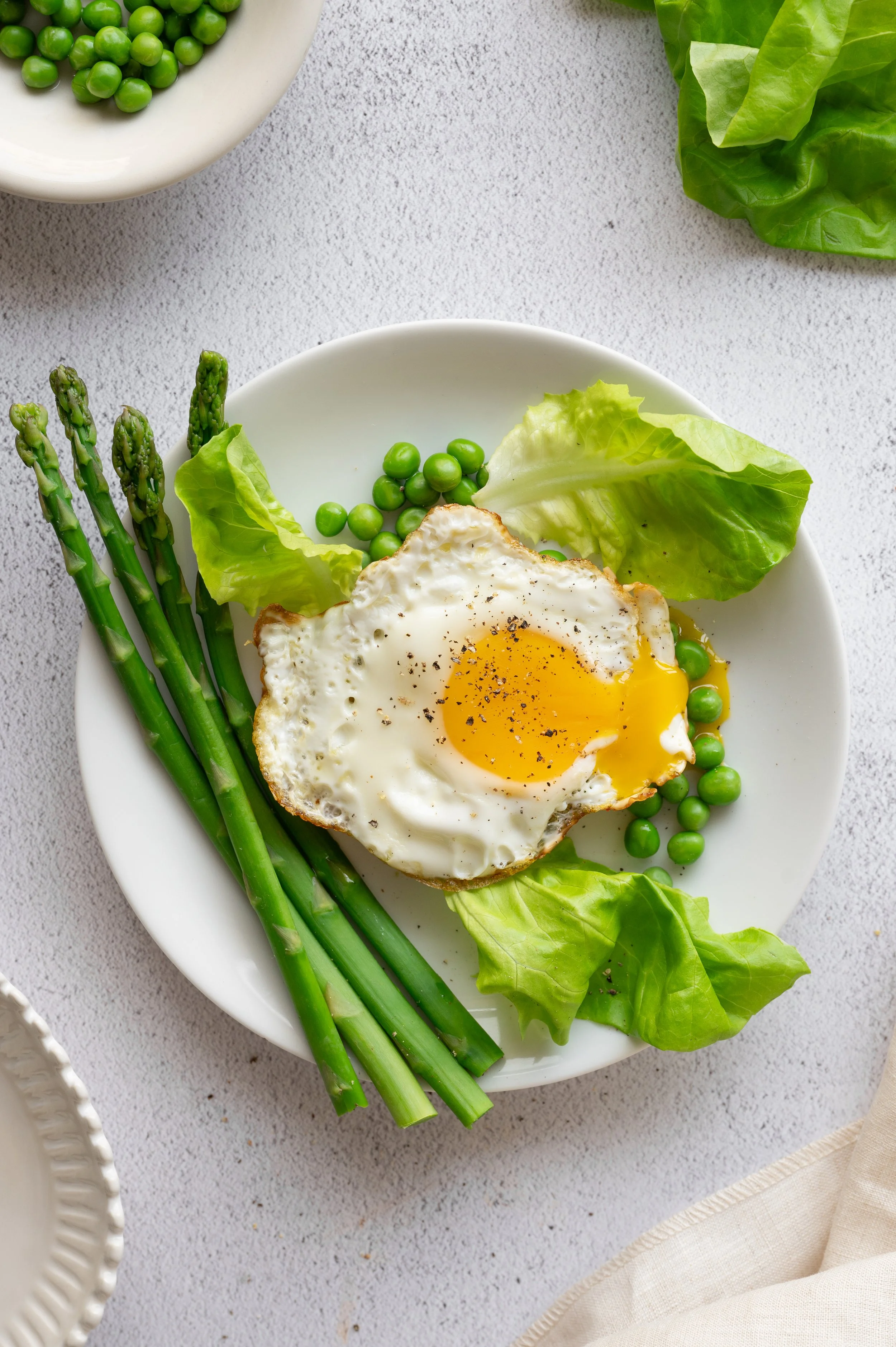 A white plate with a fried egg, green peas, lettuce, and asparagus on a white textured surface.