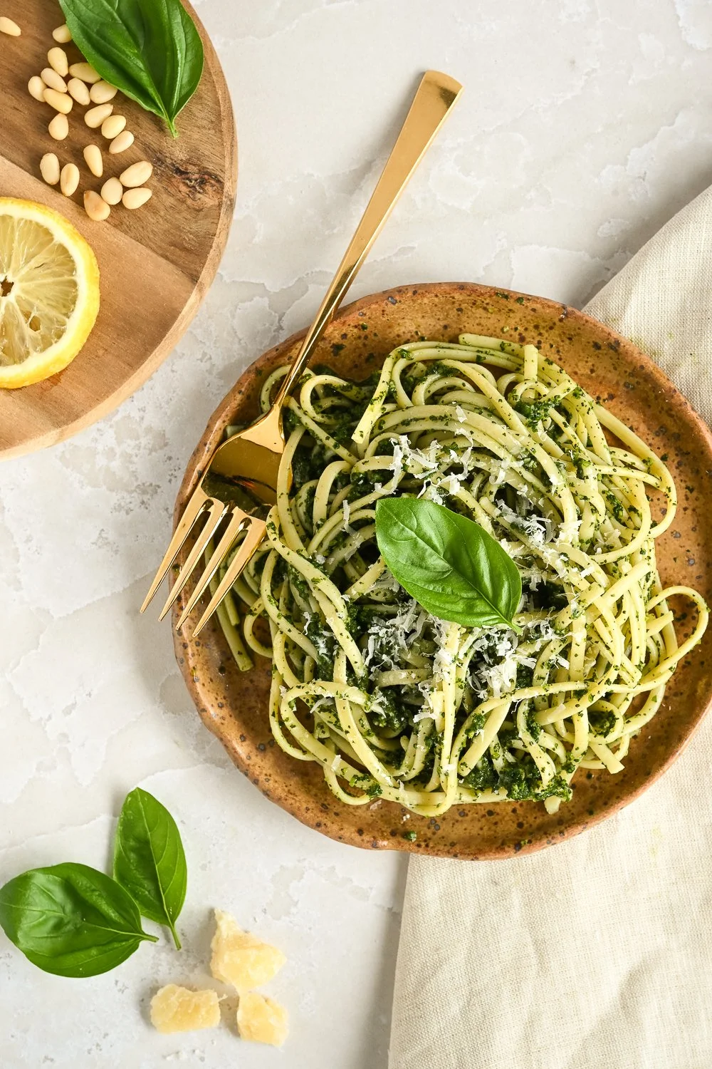 A bowl of spaghetti pasta with pesto sauce topped with a basil leaf and grated cheese, on a light-colored surface with a fork placed in the bowl. Nearby are fresh basil leaves, a lemon slice, pine nuts, and a block of Parmesan cheese.