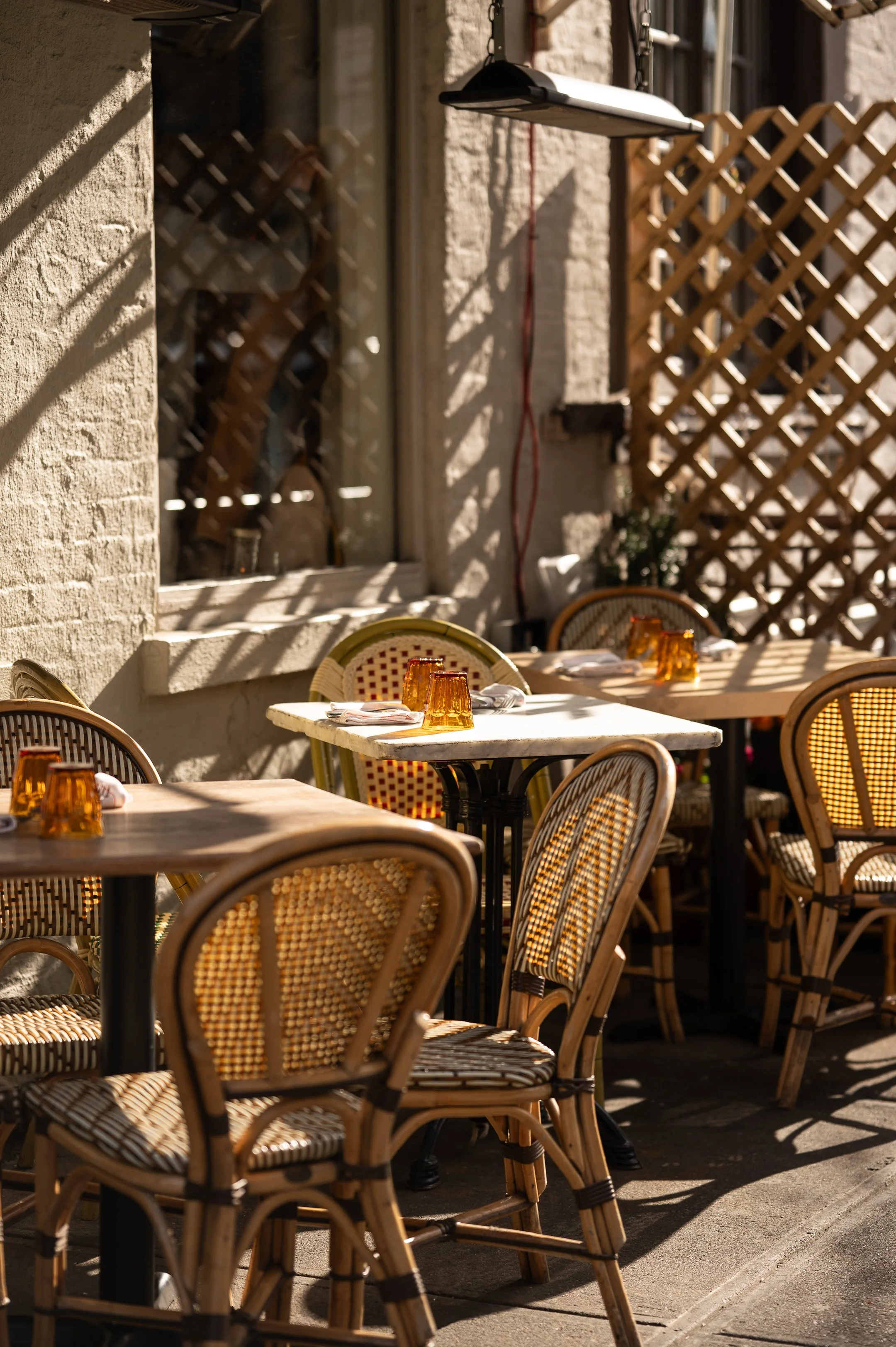 Empty outdoor restaurant patio with wicker chairs around tables, sunlight casting shadows, and a lattice fence in the background.