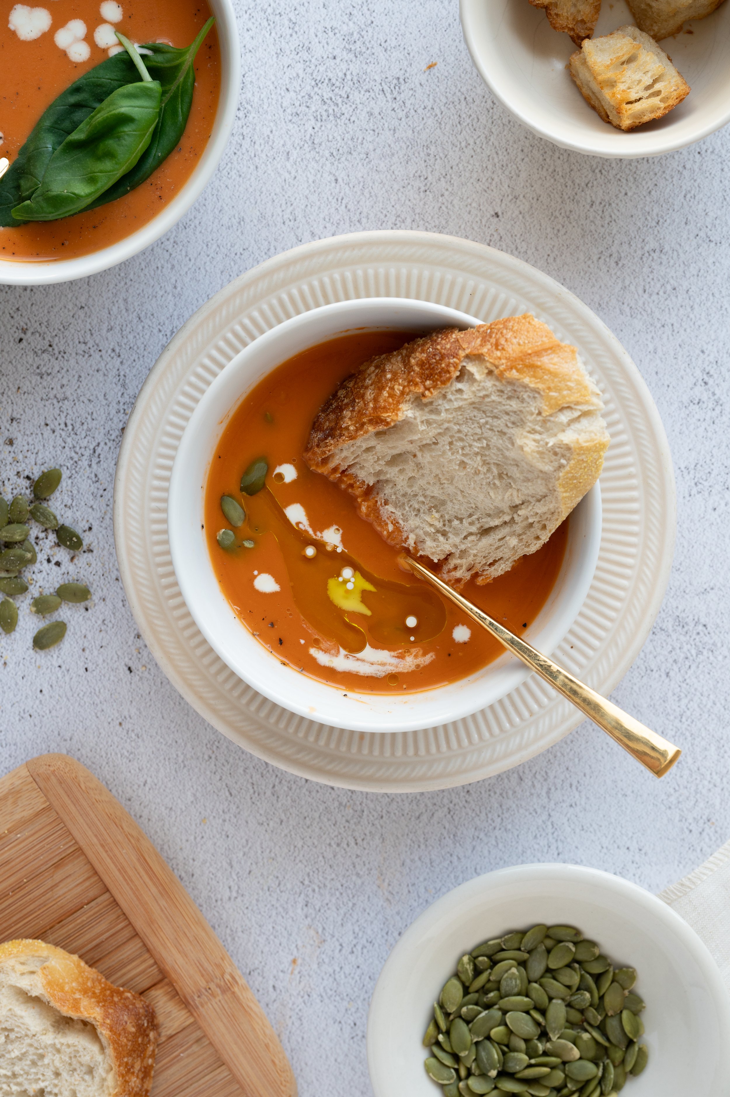 Bowls of tomato soup garnished with fresh basil leaves, pumpkin seeds, and a drizzle of oil, along with slices of bread and croutons on a white surface.