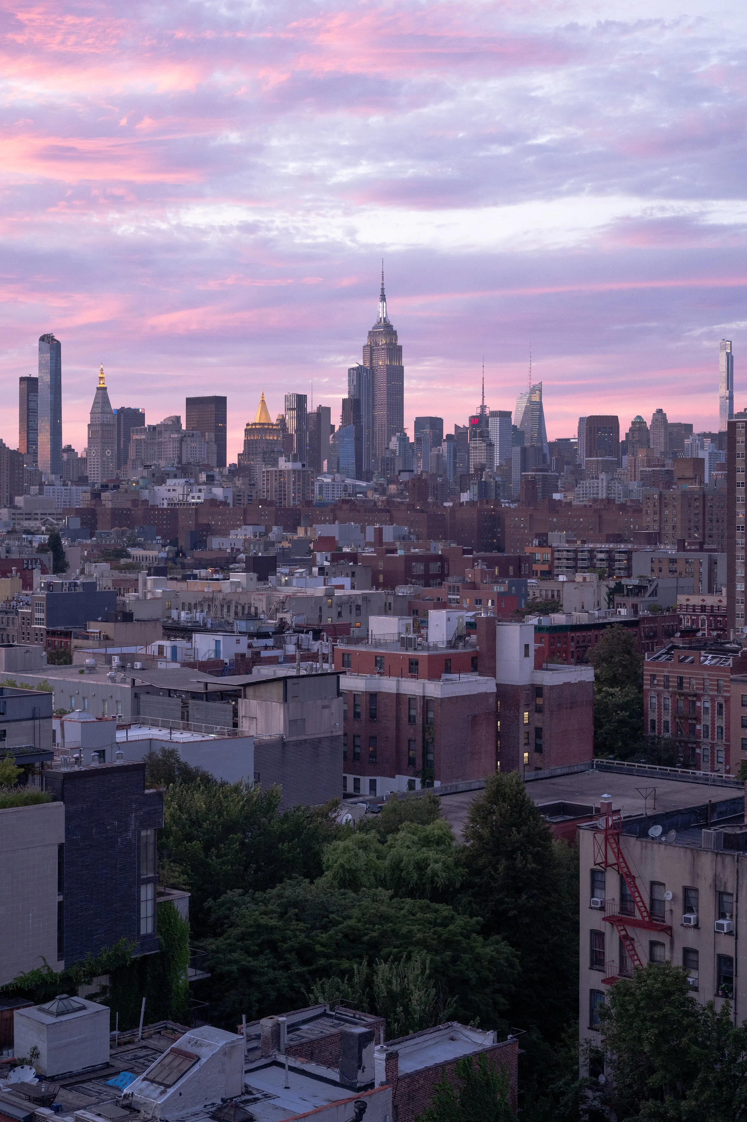 A city skyline at dusk with the Empire State Building in the center, surrounded by other skyscrapers and residential buildings, under a sky with pink and purple clouds.