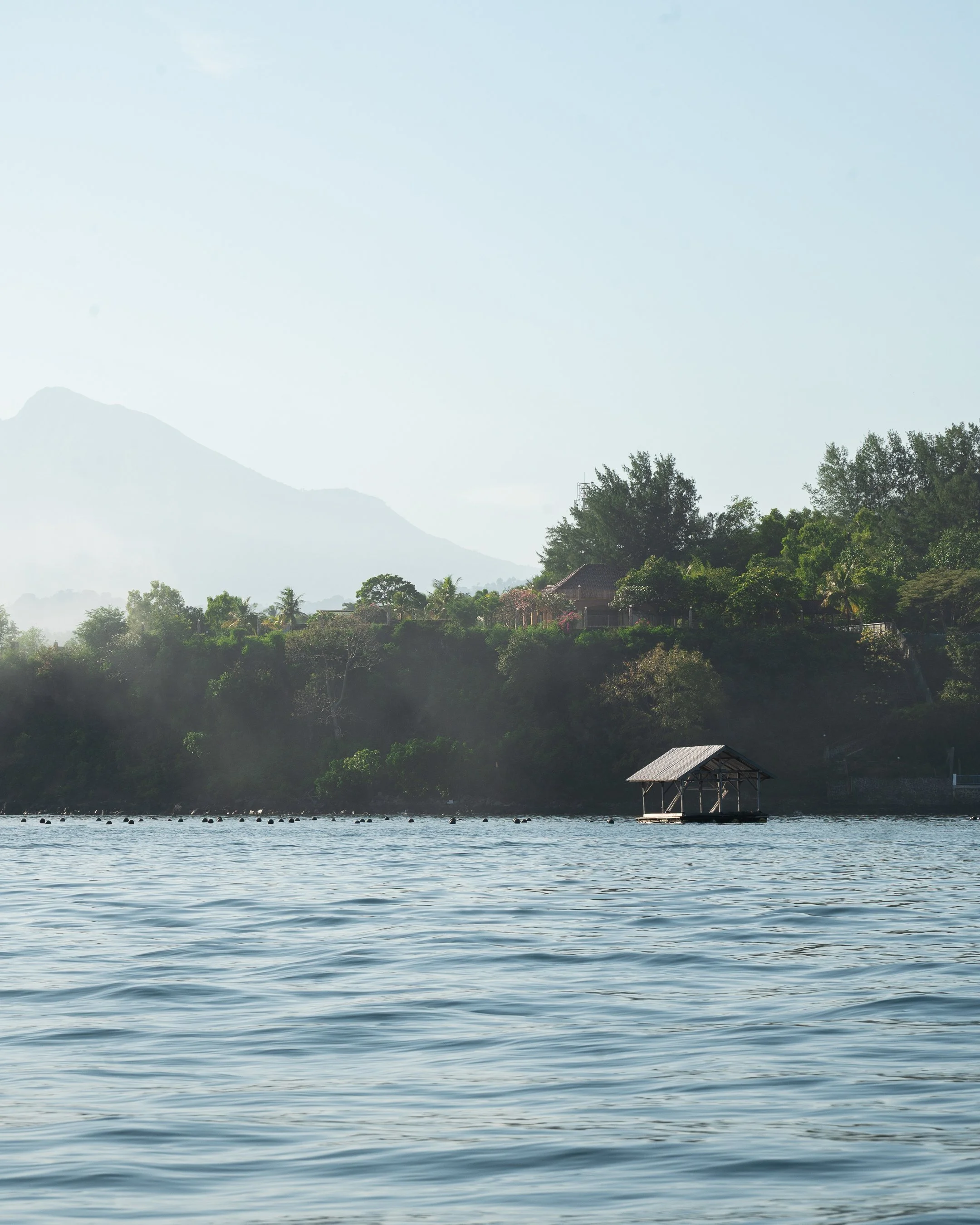 A calm body of water with small waves, a floating structure with a roof on the water, a tree-lined shoreline with houses, and misty mountains in the background under a partly cloudy sky.