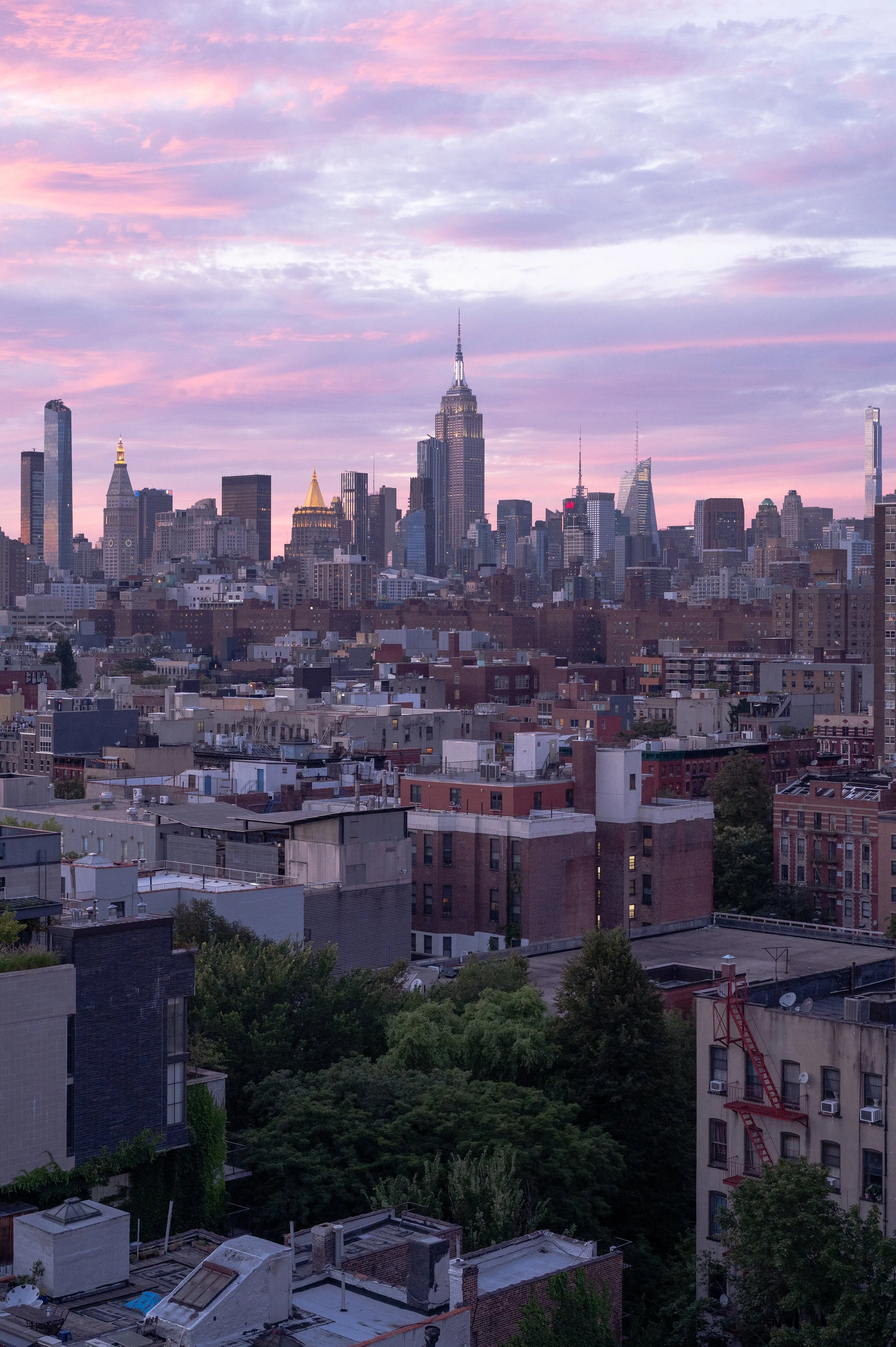 City skyline of New York City at sunset with tall skyscrapers, including the Empire State Building, under a colorful pink and purple sky, with residential and commercial buildings in the foreground.