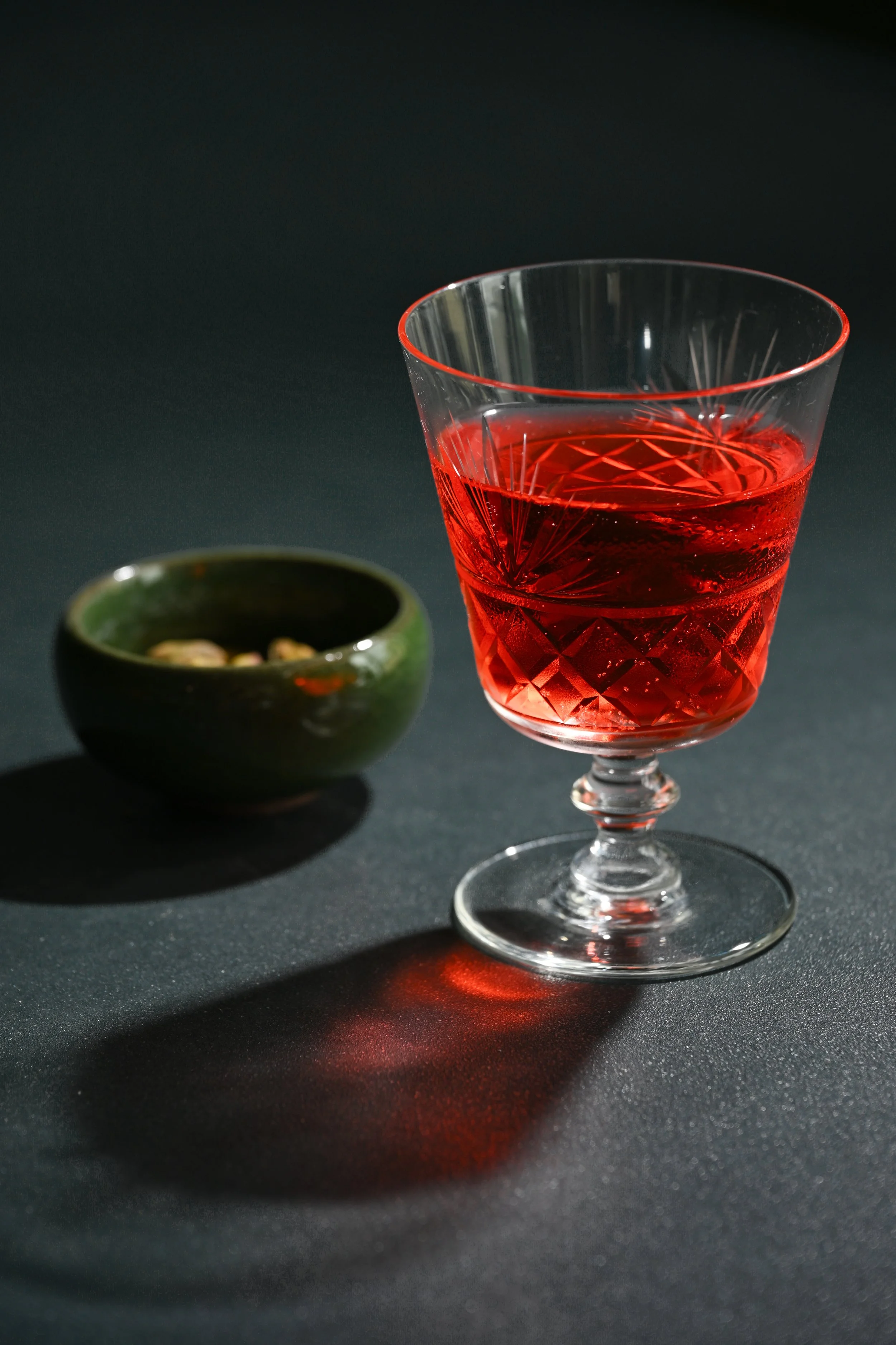 A glass of red liquid on a black surface with a small green bowl filled with nuts next to it, against a dark background.