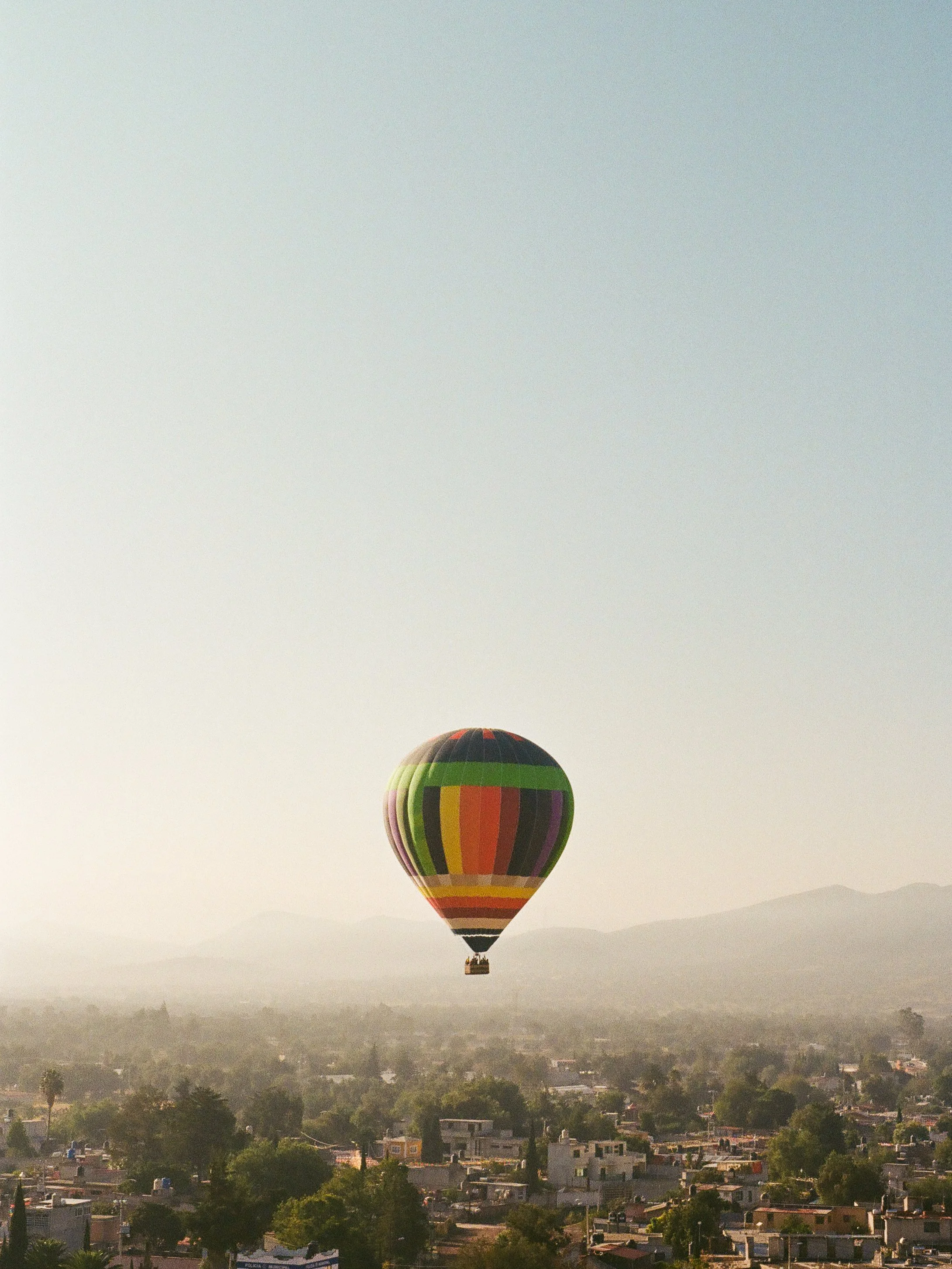 Colorful hot air balloon floating above a city with trees and buildings in the background.