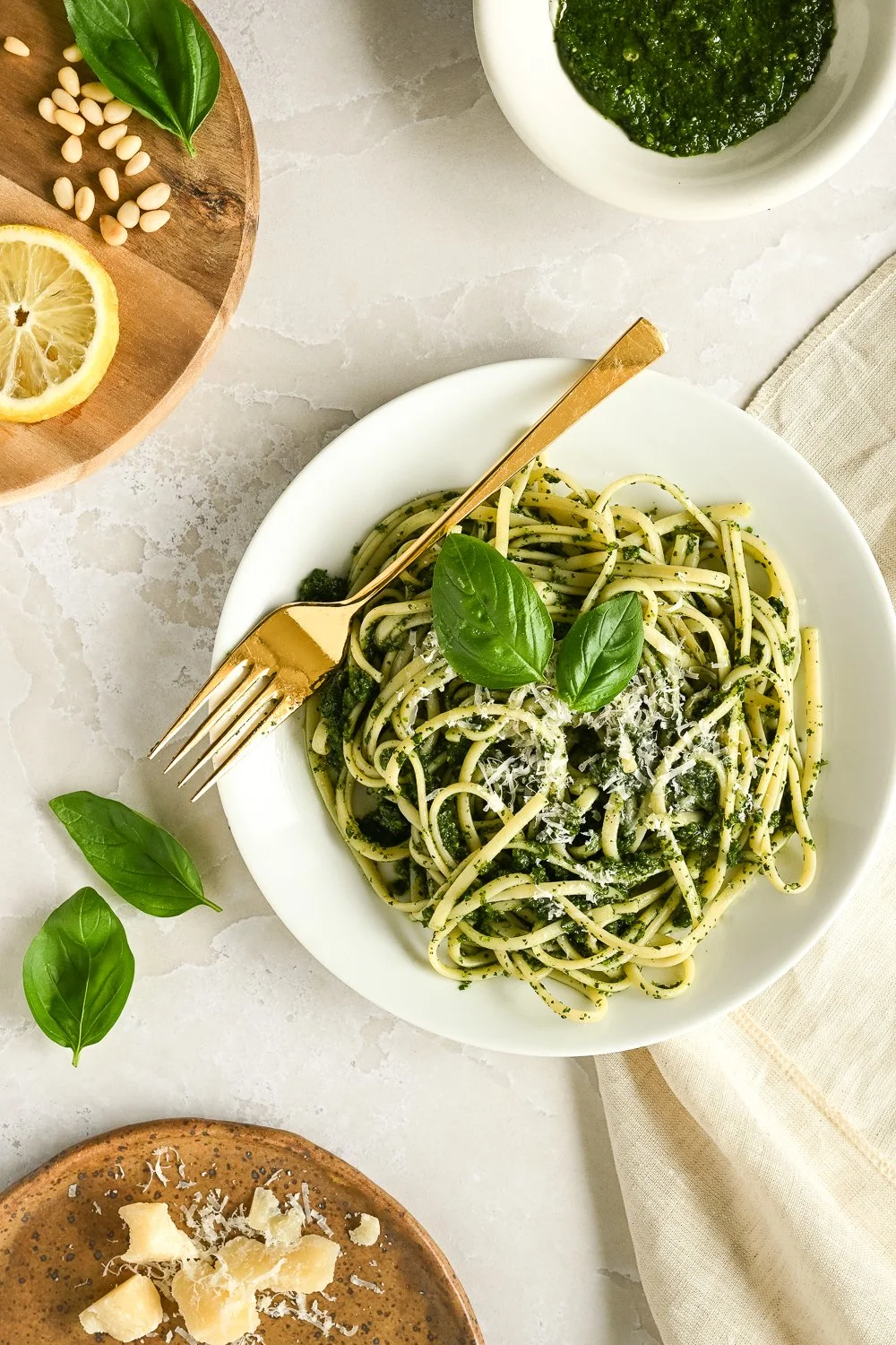 A plate of spaghetti with pesto sauce, topped with grated cheese and basil leaves, on a white plate. Near the plate are a fork and fresh basil leaves. To the top right, a small bowl of pesto. To the top left, a wooden plate with pine nuts, a lemon sl