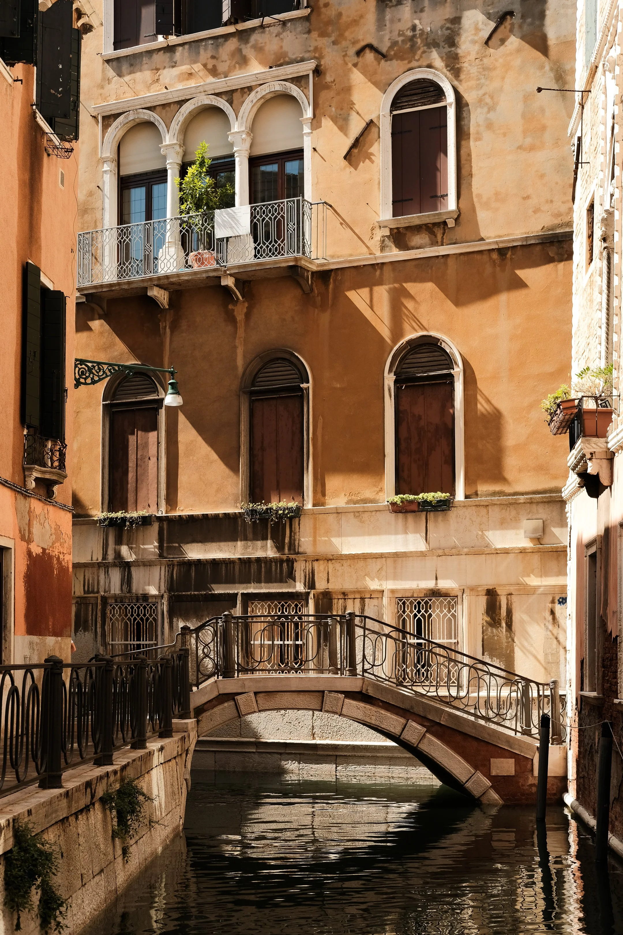 A narrow canal with a small stone arched bridge and old buildings with shuttered windows on either side. The buildings are weathered with a warm, faded yellow and orange stucco, and some windows have plants and curtains. A stone railing runs along th