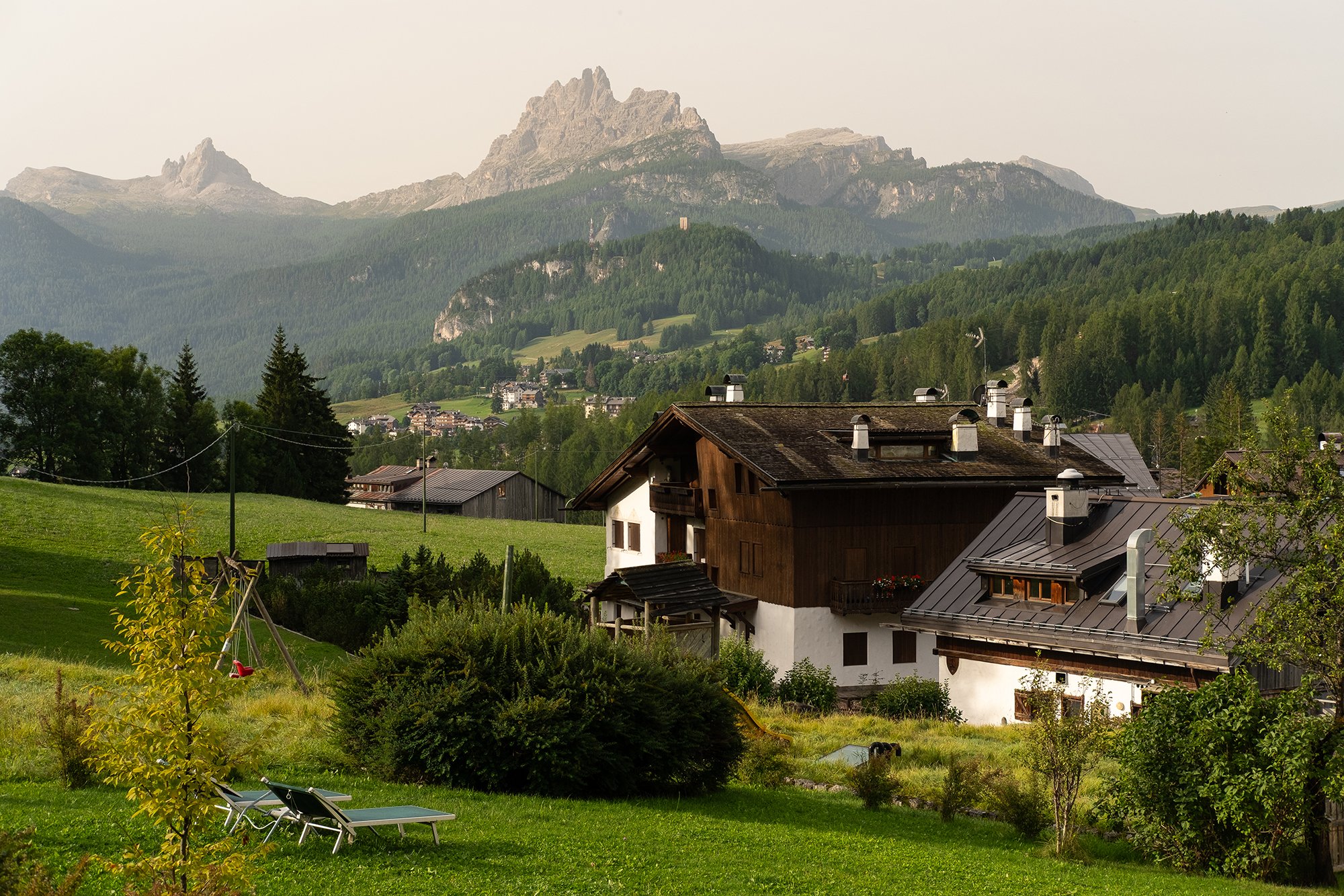A scenic view of a small village with houses and greenery in front of a mountain range with rocky peaks.