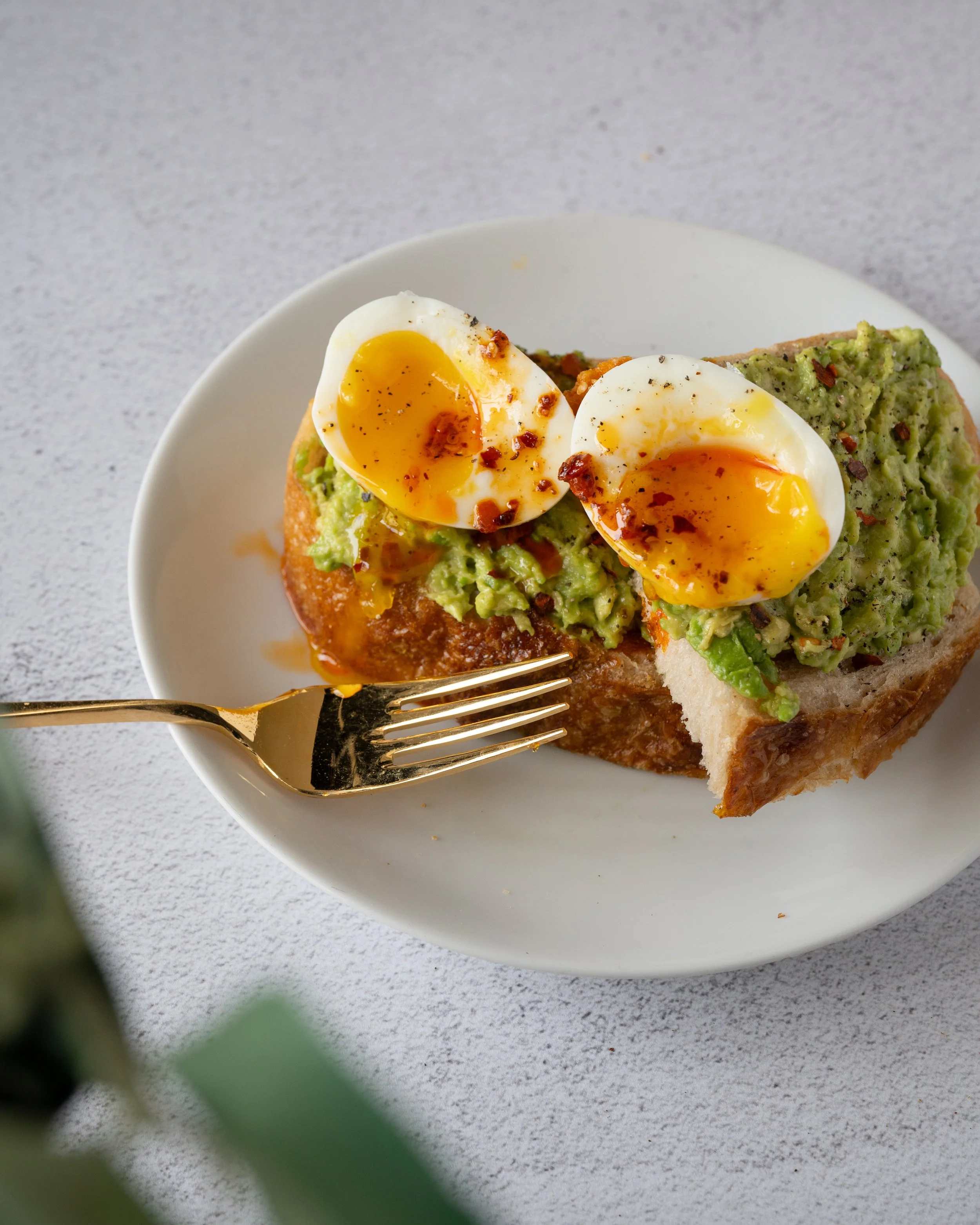 Close-up of a slice of bread topped with mashed avocado and two soft-boiled eggs, seasoned with black pepper and chili flakes, on a white plate.