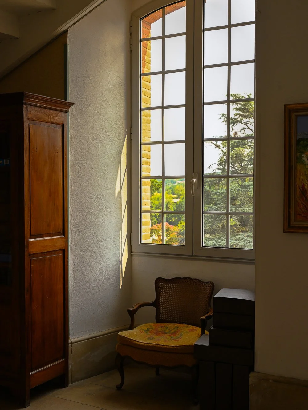 Sunlight streaming through a large window, illuminating a vintage chair with floral upholstery and a small stack of black boxes nearby, inside a room with a wooden cabinet and framed artwork.