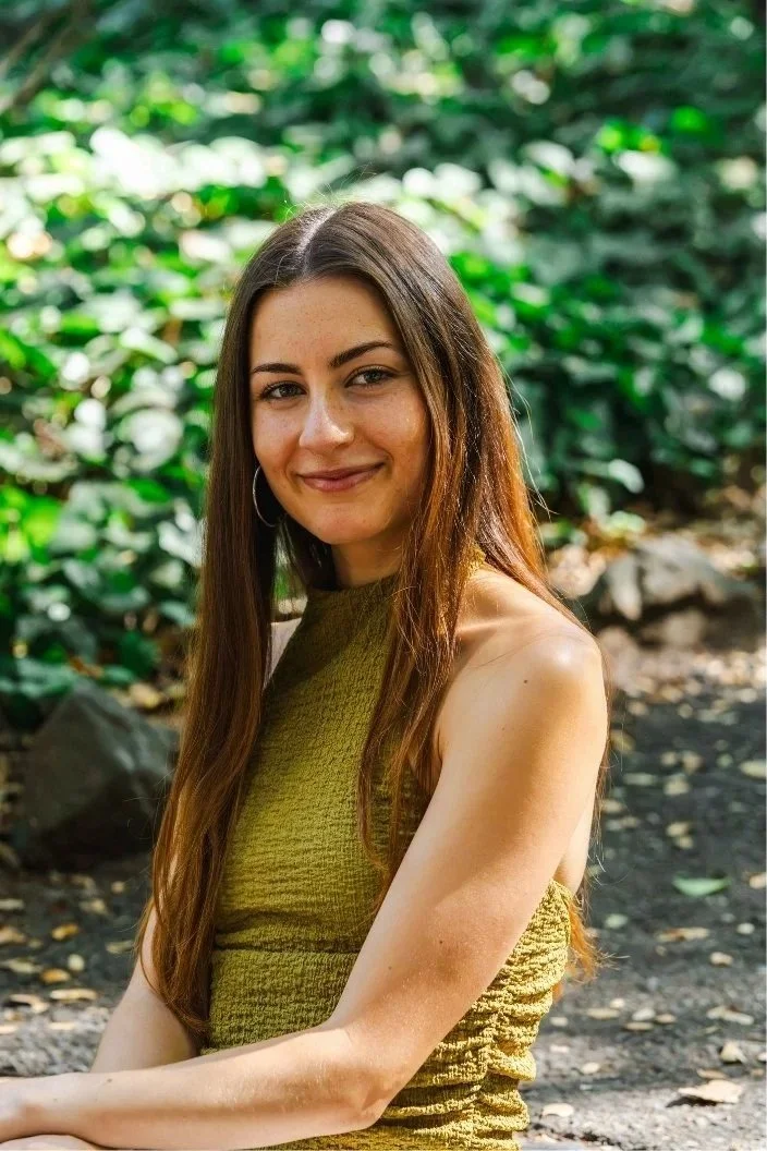 A young woman with long brown hair, wearing hoop earrings and a sleeveless green top, smiling outdoors with a background of lush green leaves.