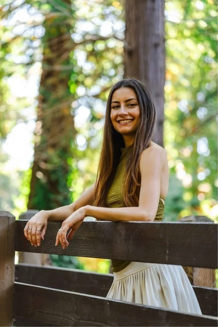 A young woman with long brown hair smiling while leaning on a wooden fence outdoors with trees and sunlight in the background.