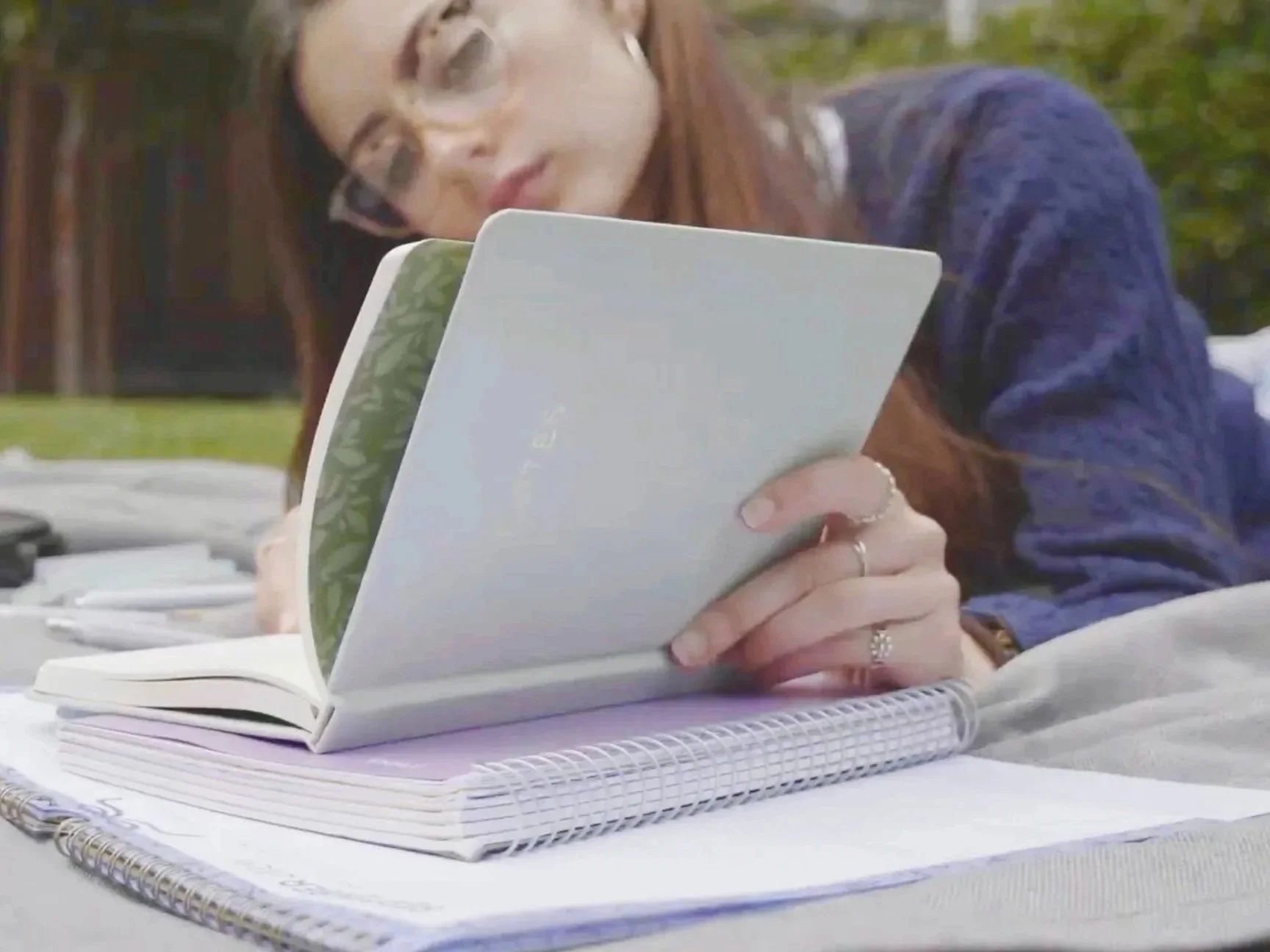 A woman with glasses and a ring on her finger reads a book outdoors at a table with notebooks and papers.